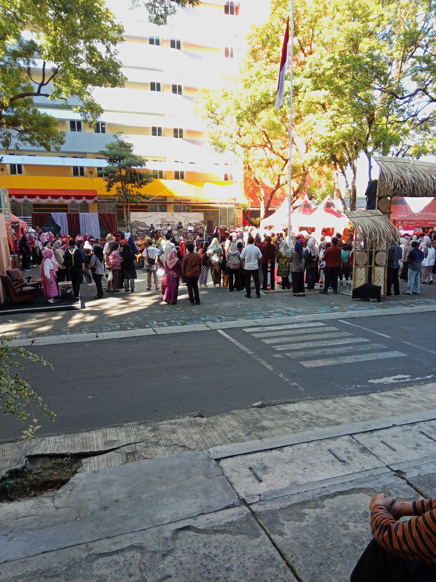 An outdoor event is taking place in front of a multi-story building with a yellow and white facade. The event appears to be a bazaar, with several tents set up. A flagpole with an Indonesian flag is visible in the background. A crowd of people, many wearing traditional Indonesian attire, are gathered, some looking towards the tents or a small stage area. A crosswalk is marked on the asphalt road in the foreground. The scene is illuminated by bright sunlight, casting shadows on the ground. The location is implied to be in Malang, Indonesia, based on the provided context, but this is not explicitly visible in the image itself.