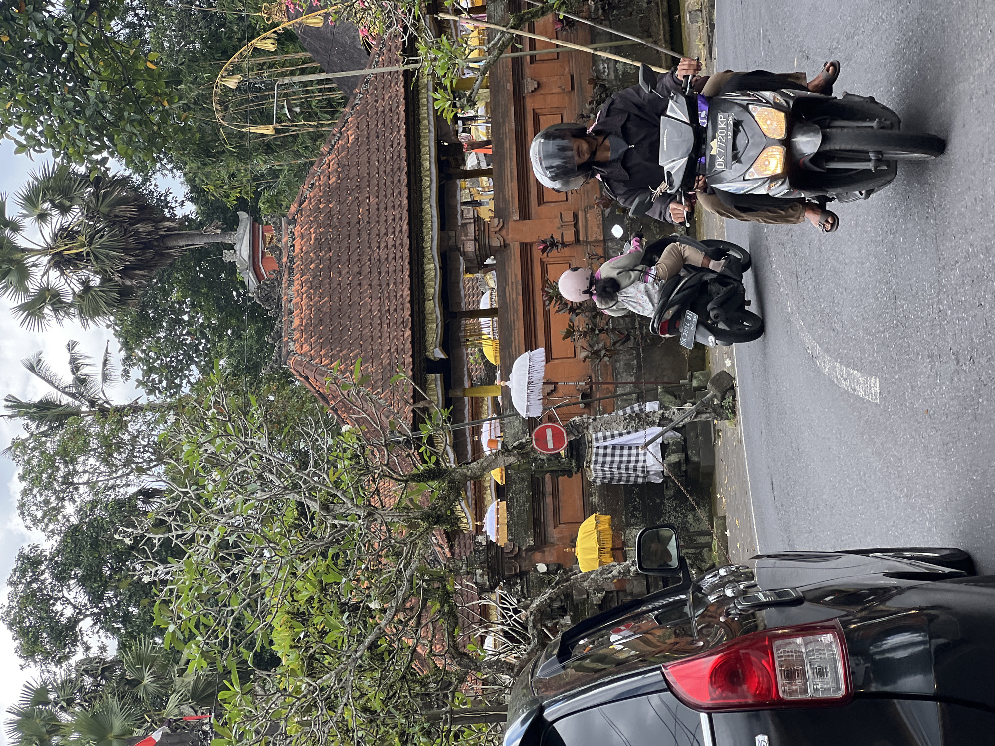 In Ubud District, Indonesia, on a cloudy day, a black car is parked on the left side of a paved road, facing forward. A man wearing a black jacket and helmet rides a black scooter, followed by a person wearing a pink helmet on another black scooter. Both scooters are heading away from the camera. Behind the scooters, a traditional Balinese building with a red-tiled roof and intricate stone carvings is visible. The building is adorned with yellow and white umbrellas. Lush green trees and palm trees surround the scene, creating a tropical atmosphere. A traffic sign with a red circle and a horizontal bar is also present.
