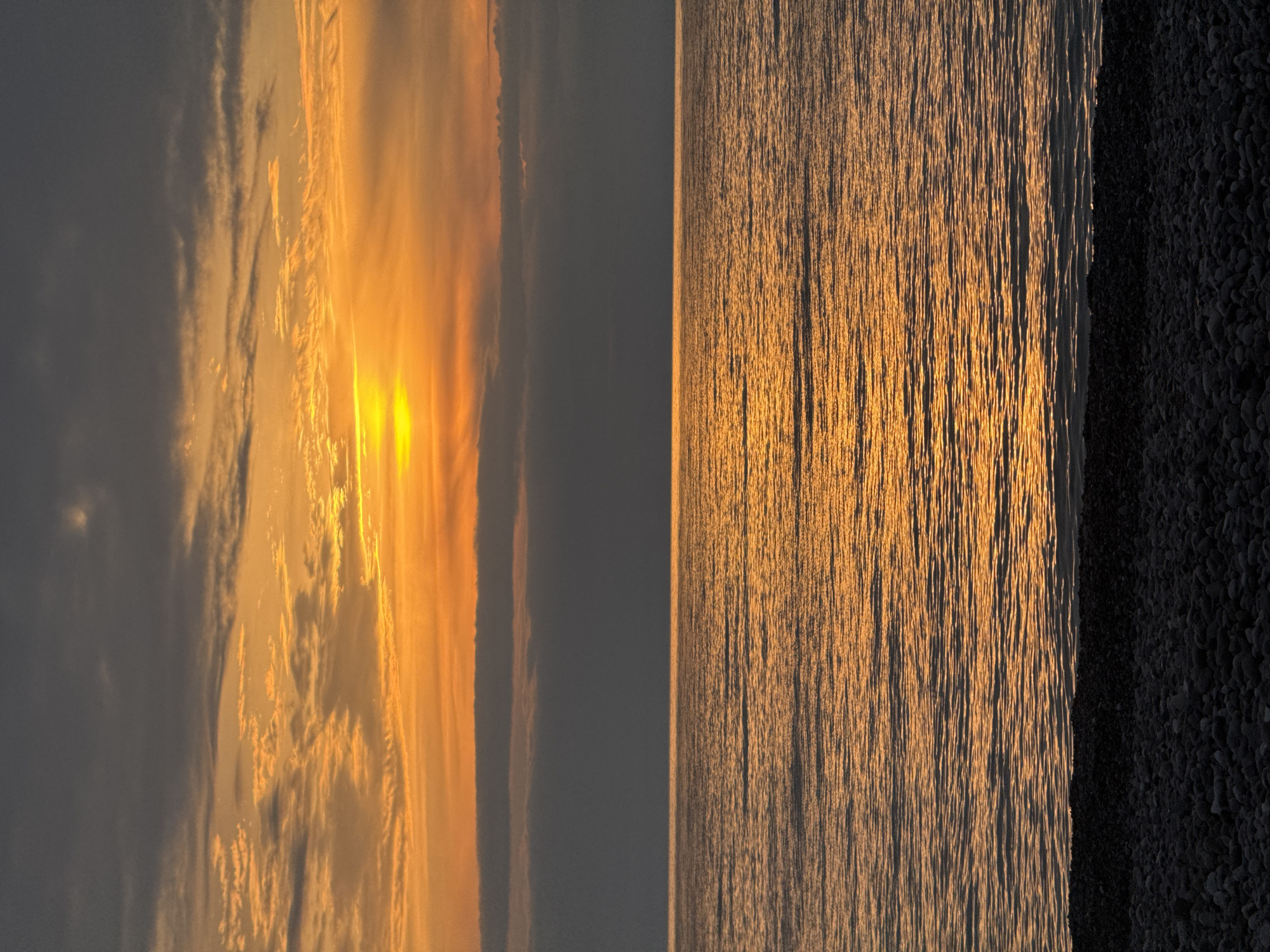 A photograph captures a sideways view of a sunset over the Black Sea in Batumi, Georgia. The sky and water are split into two sections. The sky contains clouds in shades of orange and gray, with the sun peeking through a break in the clouds. The water reflects the orange glow of the sunset. The beach is rocky.
