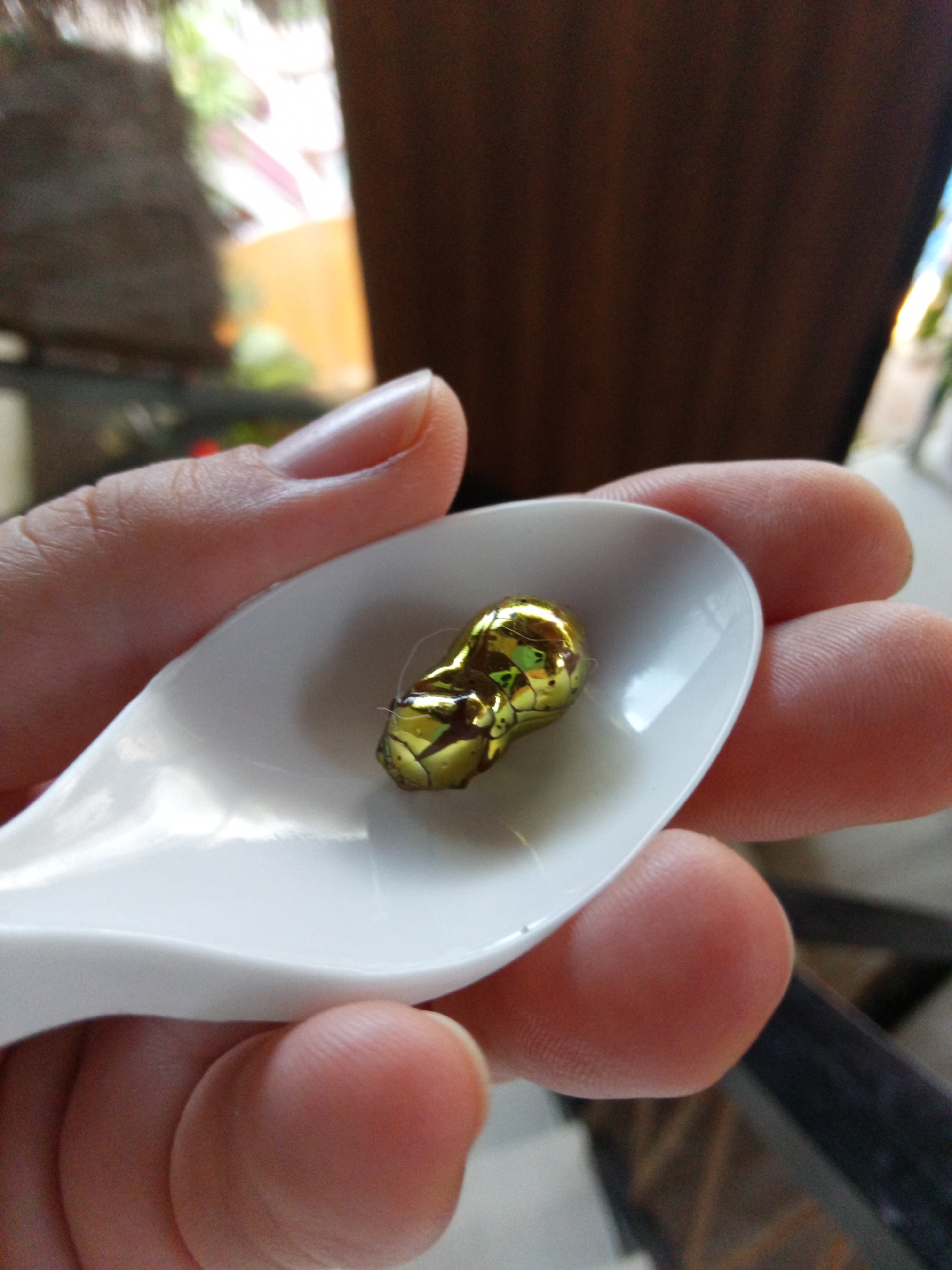 A hand holds a white plastic spoon. In the spoon is a small, gold-wrapped chocolate, possibly a Reese's Peanut Butter Cup. The background is blurred, showing hints of a building and greenery. The image was taken in Bac Natural Heritage Area Trang, Vietnam.