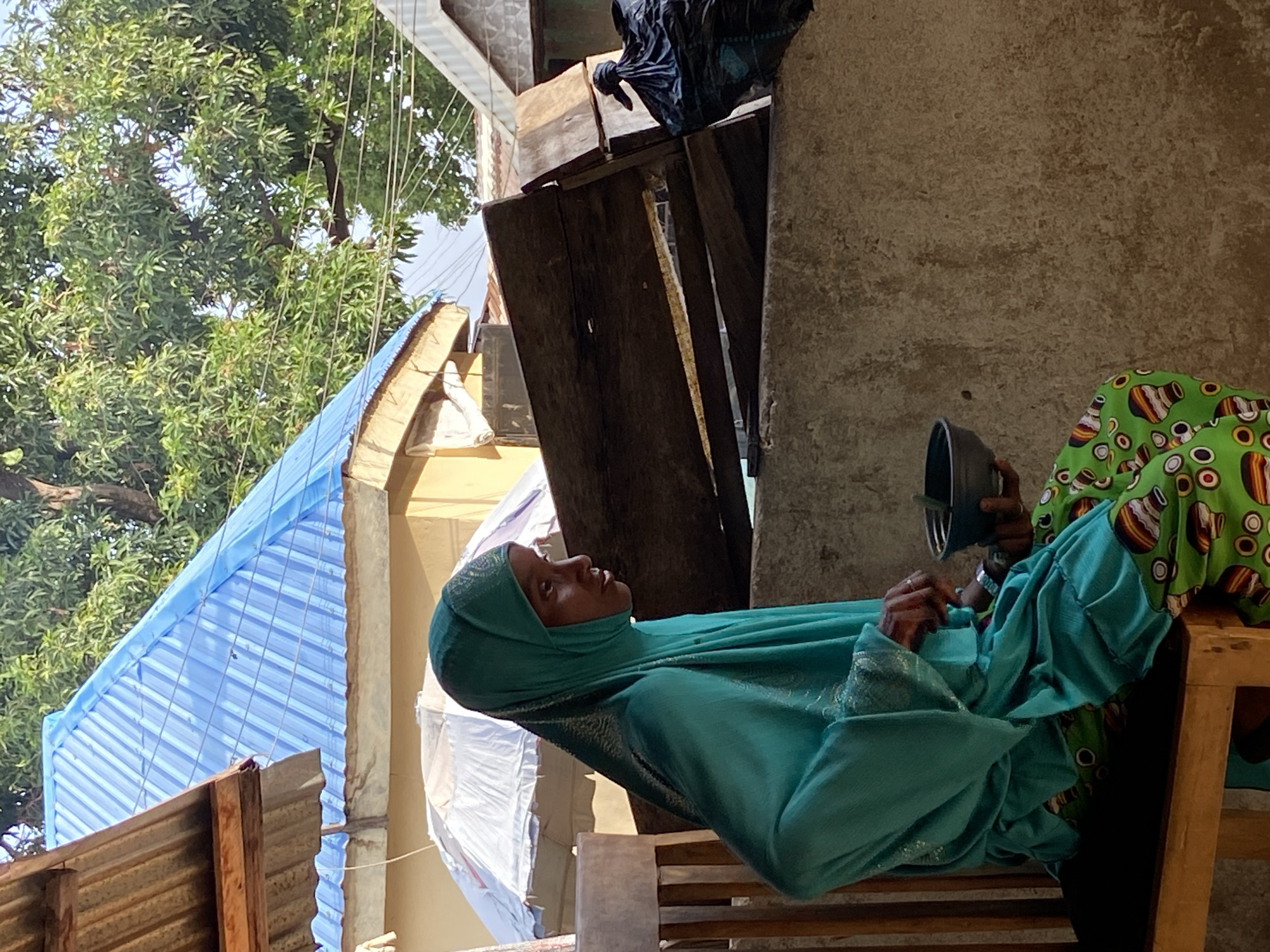 An older African woman wearing a light blue headscarf and a bright green patterned garment is seated on a wooden bench. She is holding a dark-colored circular object, possibly a bowl or container. The woman appears to be resting or sitting outdoors. The background includes a building with a light blue corrugated metal roof, a tree with lush green foliage, and a concrete wall or structure. The overall scene suggests a residential area in Mararaba, Nigeria.