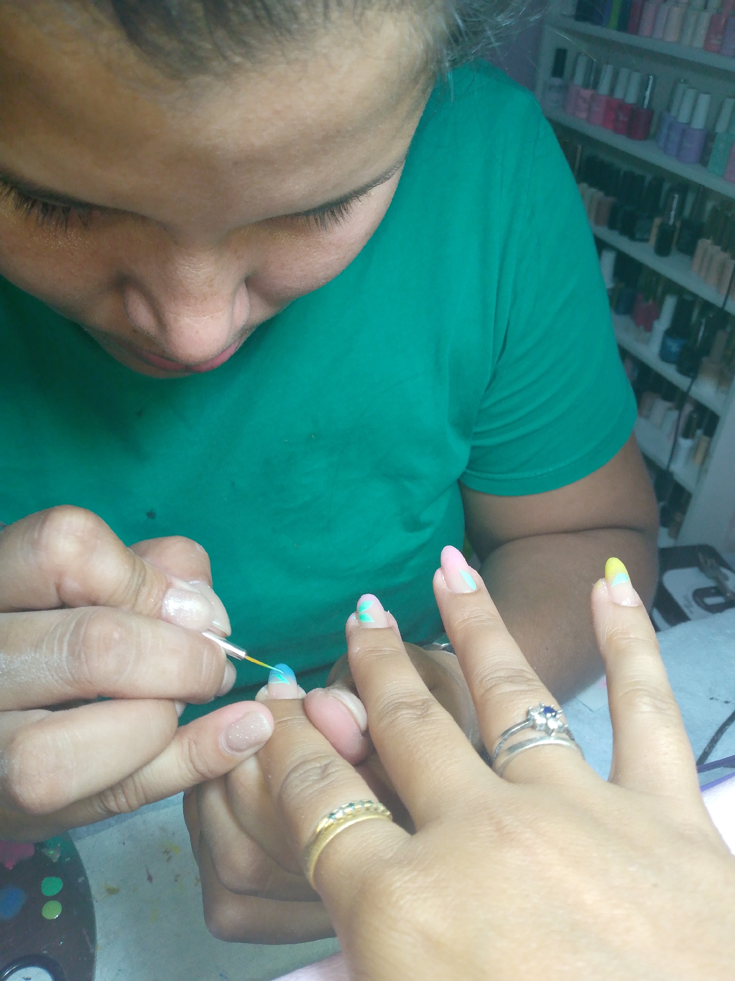 The photograph shows a woman in a green shirt performing a manicure. A manicurist is using a fine-tipped brush to apply light blue nail polish to the nail of a client's hand. The client is wearing two rings, one gold and one silver. In the background, a shelf is visible with numerous bottles of nail polish. The scene appears to be taking place in a nail salon in Tinaquillo, Venezuela, based on the user's description.