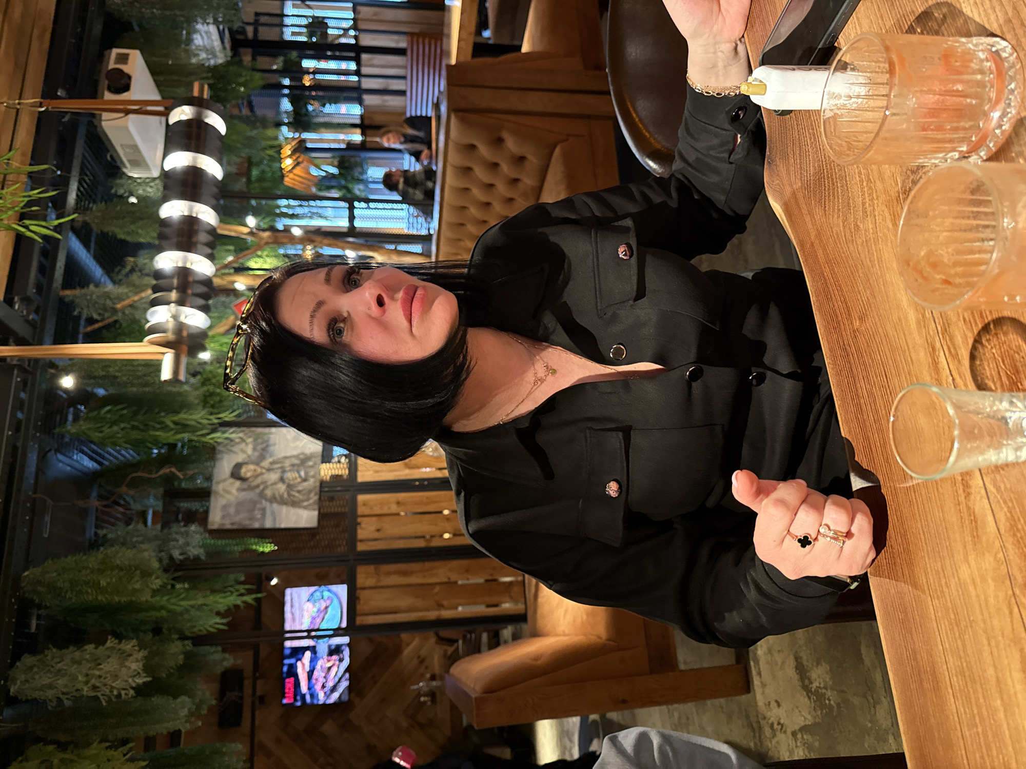 This image captures a woman seated at a wooden table in what appears to be a modern restaurant or bar, likely during the evening in Malakhovka, Russia. The woman, with short dark hair and glasses resting on her head, looks towards the camera with a neutral to slightly serious expression, revealing a nose piercing. She is wearing a black collared top or jacket with button detailing and a delicate gold chain necklace. Her left hand, adorned with multiple rings including one with a black clover design and another with a pearl, rests on the table.
On the table in front of her are several empty or partially filled ribbed glasses, one containing a white and gold dispenser-like object. The background reveals the restaurant's interior decor, which includes artificial green plants or shrubs, modern cylindrical light fixtures emitting soft light, and cushioned booth seating. Further back, out-of-focus figures of other patrons are visible. The walls feature wooden paneling, decorative metal grating, and what seem to be framed paintings or artwork. Two television screens are also visible, displaying colorful abstract images, with the word "BURGER" faintly discernible in red text on one. The overall atmosphere is dim and ambient, suggesting a relaxed dining or socializing environment.