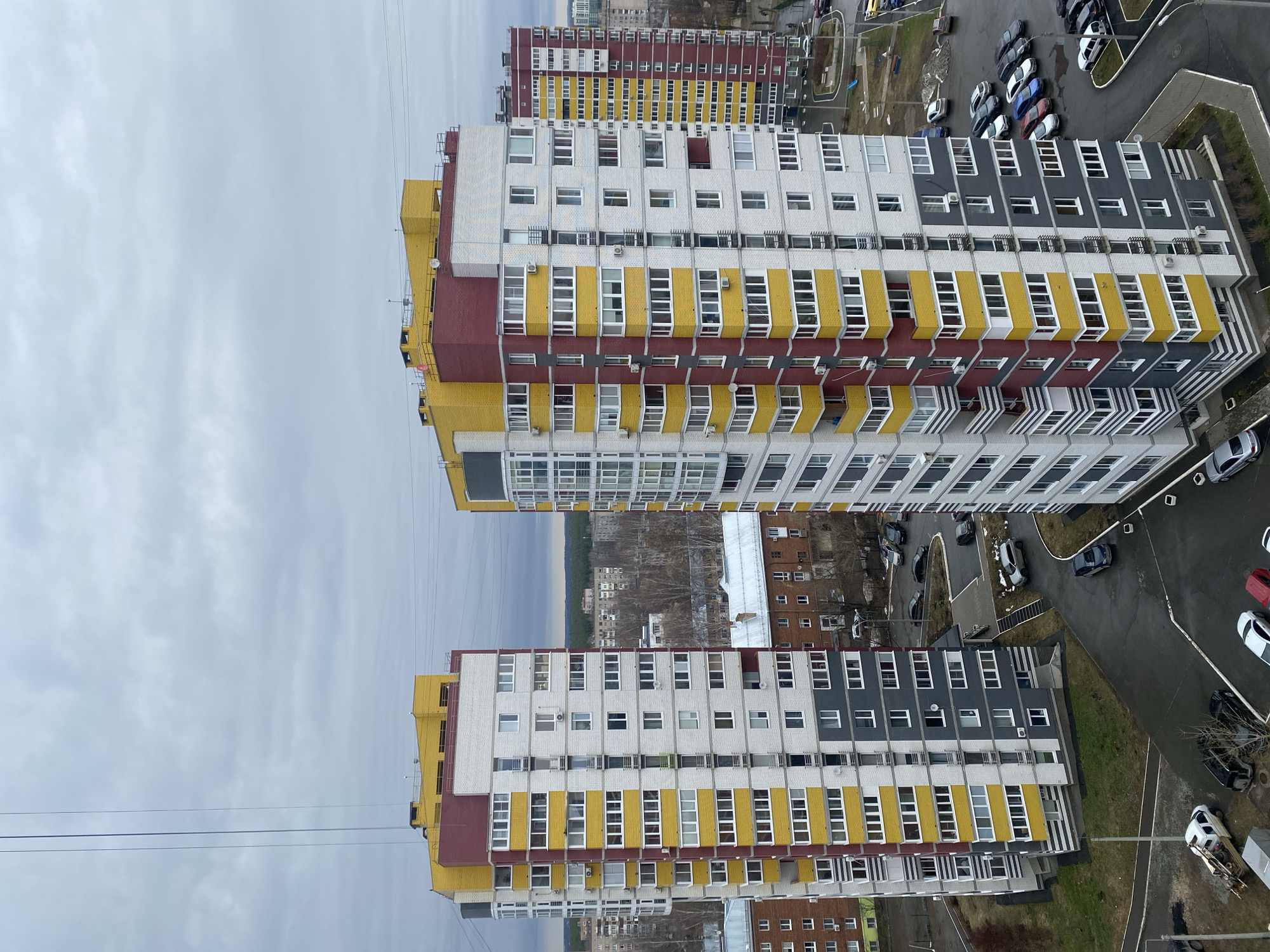 The image, taken from a high vantage point, shows several apartment buildings in Izhevsk, Russia. The buildings have a recurring design with white, yellow, gray, and maroon accents. They are oriented in a way that showcases their rooftops and sides. Cars are parked near the buildings, and a street is visible. The environment suggests an urban setting. The sky is overcast.