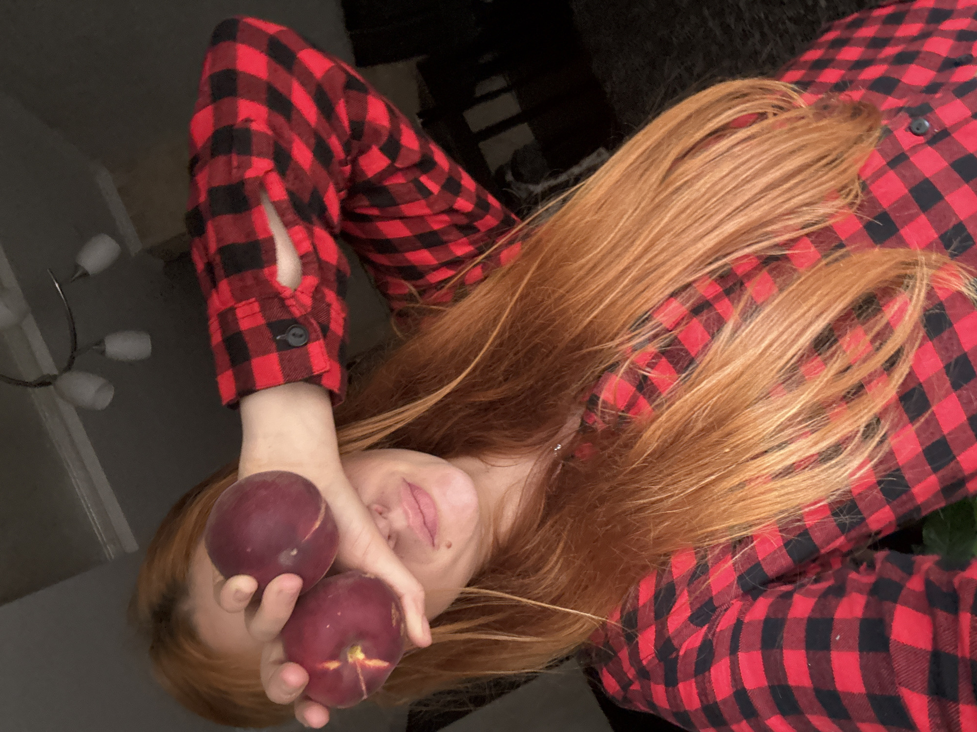 An angled, close-up indoor shot features a person, likely female, with long, flowing reddish-brown hair cascading over a red and black buffalo plaid shirt. The person's head is tilted, with only a portion of their face visible, including the chin, mouth, and parts of the nose and left eye area. Her lips are slightly parted, suggesting a relaxed or subtly smiling expression, with a small mole visible on her chin.
Two dark reddish-purple fruits, resembling plums or nectarines, are held in one hand, strategically covering most of the person's eyes and upper face. The plaid shirt has black buttons on the placket and a sleeve, with a distinctive slit opening near the cuff of the left sleeve. The background is dimly lit and largely out of focus, appearing to be an interior setting. A blurry modern light fixture with multiple white shades is partially visible in the upper left corner of the frame. The overall mood is casual and somewhat playful. The image was taken in Budva, Montenegro. There is no visible text.
