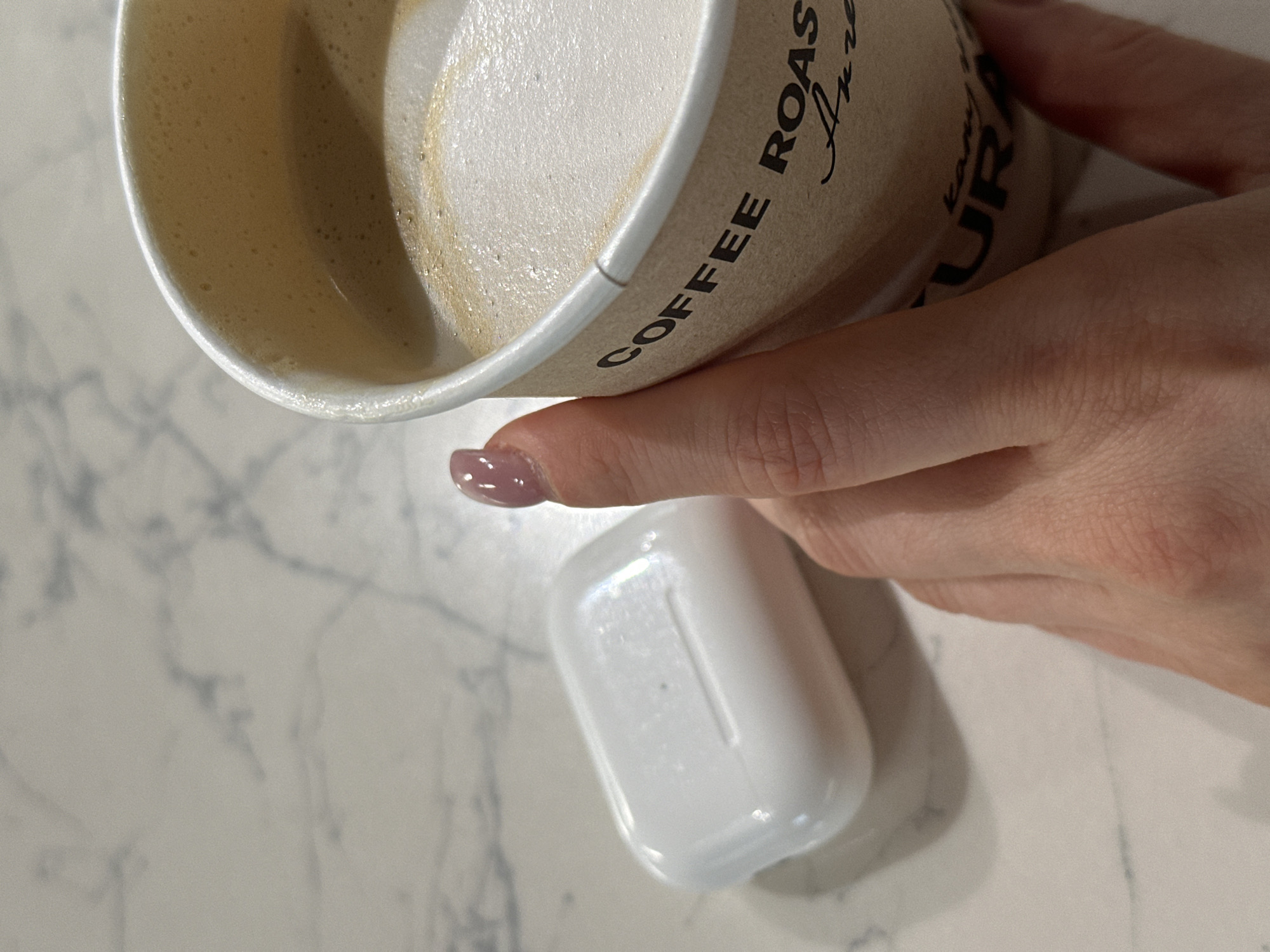 A close-up shot depicts a hand holding a paper coffee cup next to a white earbud case on a light-colored marble surface. The coffee cup is filled with a latte. The text "COFFEE ROAST" is visible on the cup. The hand's nail is painted a mauve color. The location is Kazan, Russia.