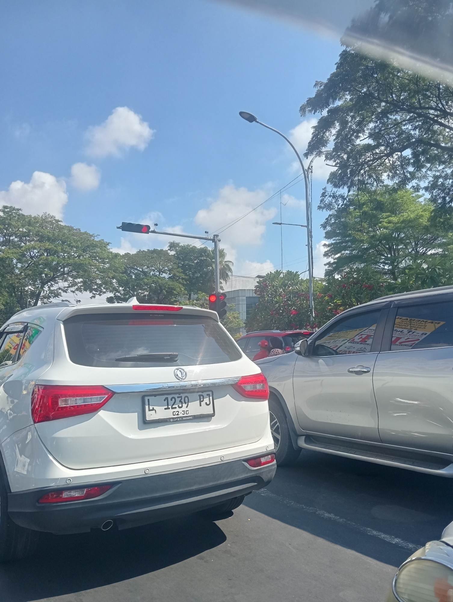 The image depicts a street scene in Surabaya, Indonesia, under a bright, partly cloudy sky. Two SUVs are prominently featured, stopped in traffic. The foreground is dominated by the rear of a white SUV, its license plate reading "L 1239 PJ". To its right, a silver SUV is also visible, its side with tinted windows and a faint reflection of yellow signage. In the background, a traffic light displays a red signal, indicating a halt in traffic flow. Trees with lush green foliage line the street, and a modern building is partially visible behind them. The overall impression is that of a typical urban traffic situation on a sunny day in Surabaya.