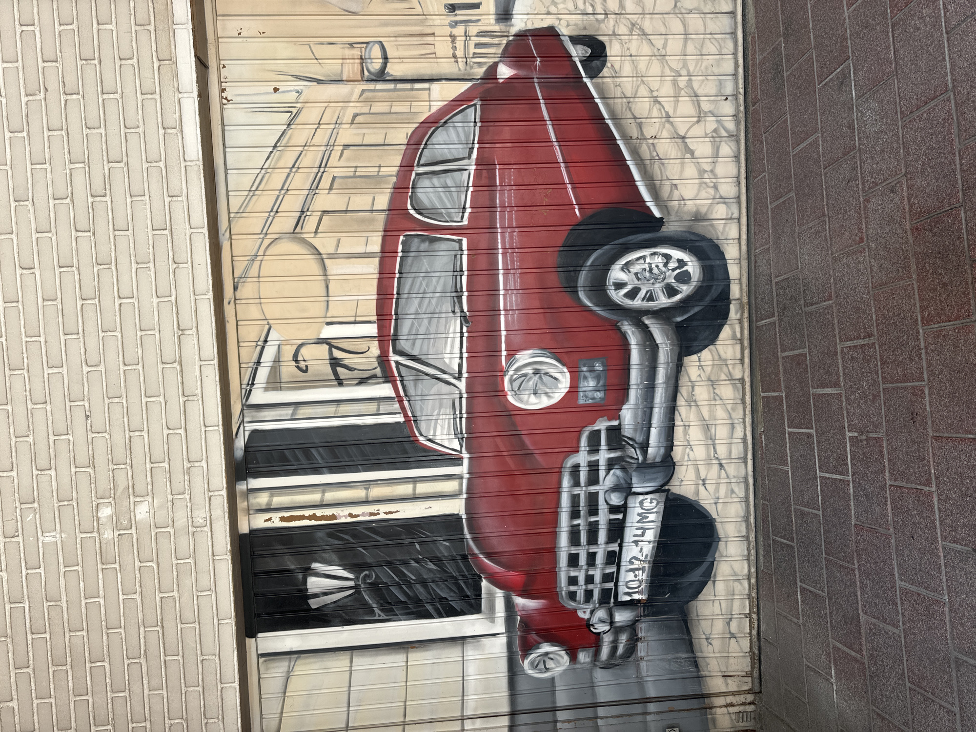 The image shows a mural painted on a ribbed metal garage door, depicting a vintage red car parked on a cobblestone street in front of a building. The car is a classic American model, possibly from the 1950s, with chrome accents and a distinctive grille. The building in the background is rendered in a monochromatic style, with windows and architectural details suggesting an urban setting. A street lamp is also visible. Above the garage door is a brick wall. The lower portion of the image shows a brick-paved sidewalk. The mural is located in the city of Murcia, Spain, specifically on a garage door on the exterior wall of the building at Calle del Ángel, 14.