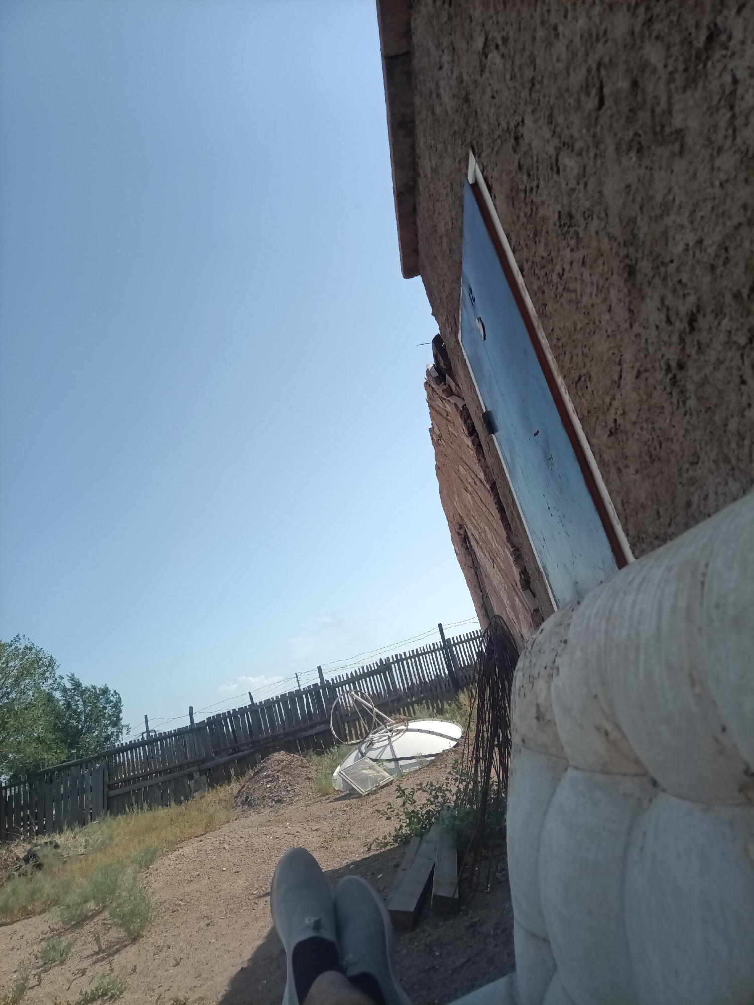 The image depicts an outdoor scene with a clear blue sky dominating the upper portion. In the foreground, the lower half of a person's legs are visible, wearing gray slip-on shoes. They are positioned on a dirt and gravel ground, with sparse dry vegetation.
To the left and middle ground, a weathered, dark wooden fence stretches across the frame. Beyond the fence, trees with green foliage are visible. A pile of dirt or earth is situated near the fence. Several discarded objects are scattered on the ground near the fence, including what appears to be part of a satellite dish and some metal scraps.
On the right side of the image, a portion of a building with a rough, textured, light brown wall is prominent. A dark blue door with a white frame is set into this wall. The building appears to be made of adobe or a similar material and shows signs of age and possibly disrepair. In the very right foreground, a large, padded, light-colored object, possibly an outdoor cushion or part of a piece of furniture, is partially visible.
The scene suggests a rural or semi-rural setting, possibly in Balqaş, Kazakhstan, given the dry vegetation and the rustic construction of the fence and building. The lighting indicates it is daytime, likely sunny. There are no other people or animals clearly visible in the image.