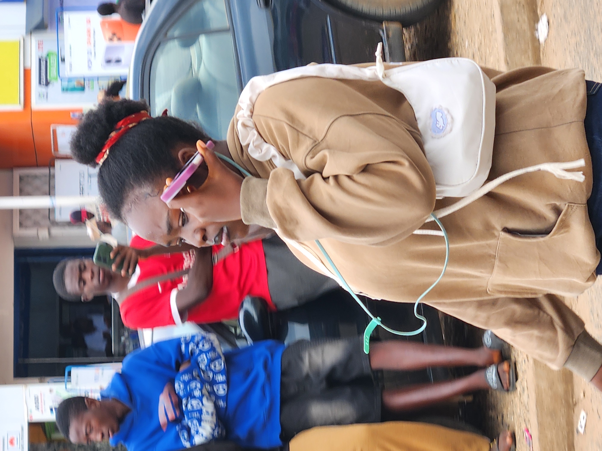 A medium shot captures a young Black woman with her hair pulled up in a bun, adorned with a red bandana, speaking on a pink cell phone in what appears to be an outdoor market or street scene in Unknown, Unknown. She is wearing a light brown hooded sweatshirt and a small white purse with a blue cloud design hangs from her shoulder. She is positioned in the foreground, slightly to the right of the frame, looking to her left. To her left, two young Black men are visible. The man closest to her wears a red t-shirt and is holding a cell phone. The man further left wears a blue hooded sweatshirt with a white design and has his arms crossed. The background includes a building with a blue awning, an orange section, and various signs, some displaying images of phones. A dark-colored car is partially visible to the right of the woman. The ground is covered in dirt and gravel. The lighting suggests it is daytime, possibly overcast.