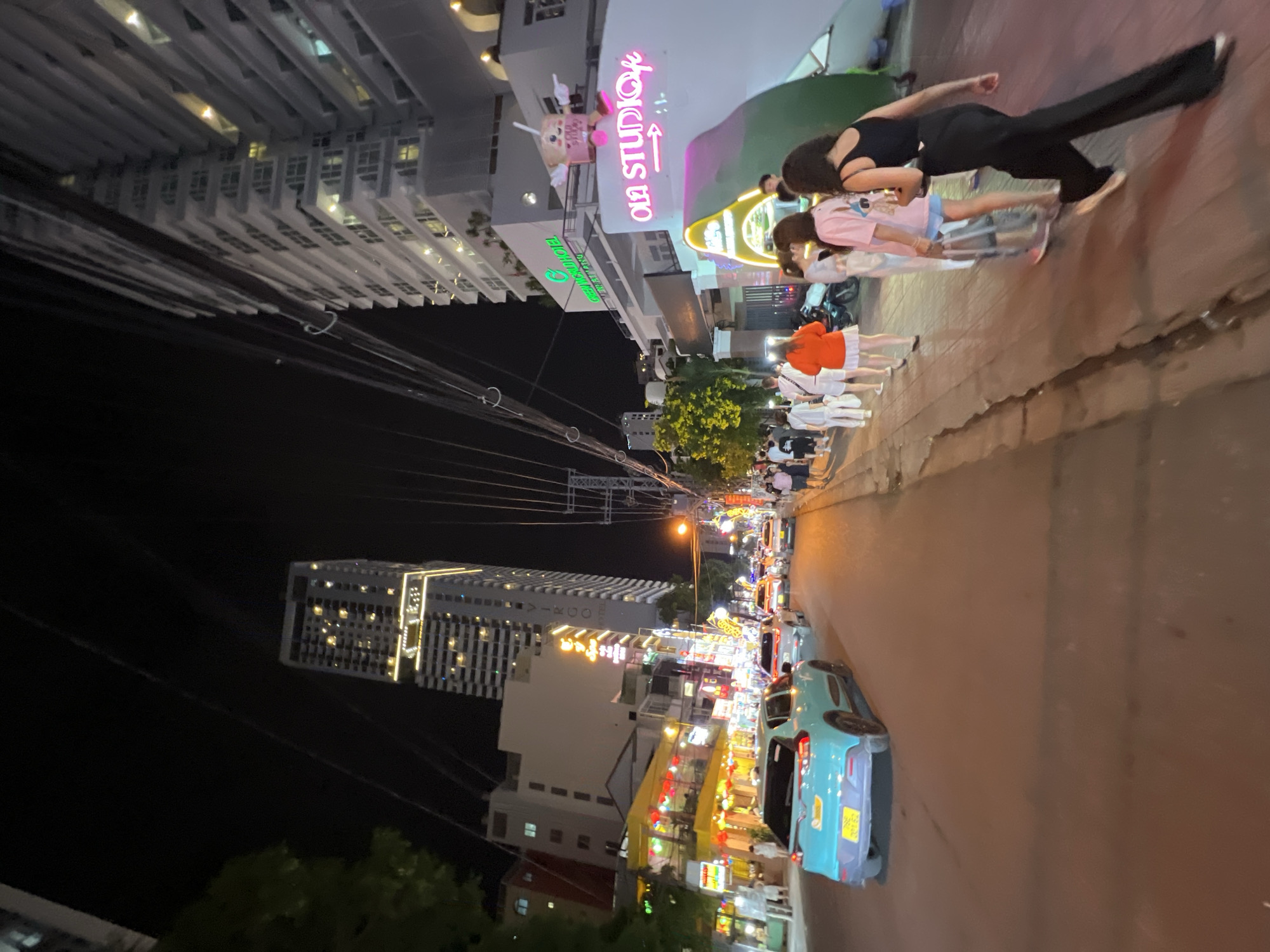 This image depicts a nighttime street scene in Nha Trang, Vietnam, specifically on Tran Phu Street, a prominent coastal boulevard known for its nightlife and hotels. The scene is illuminated by streetlights, neon signs, and the lights from buildings, creating a vibrant and active atmosphere.
In the foreground, a bright turquoise hatchback car is parked on the right side of the road, facing away from the camera. The street is paved with asphalt, and a sidewalk runs along the right side. A group of people, dressed in casual summer clothing, are walking along the sidewalk, heading towards the right of the frame. They appear to be tourists or locals enjoying an evening stroll. One woman in the foreground is wearing a black tank top and wide-leg black pants, while another person next to her is dressed in a pink t-shirt and shorts. A person in an orange shirt and white skirt is also visible.
On the right side of the image, a modern, multi-story building stands tall, with balconies and numerous lit windows. A neon sign that reads "OLA STUDIO" in pink and white lettering is prominently displayed on the building's facade, along with an arrow pointing to an entrance. A cartoonish character resembling a cup of bubble tea with arms and legs is perched above the sign. Another sign in green lettering, partially obscured by power lines, reads "GREEN WORLD HOTEL".
Further down the street, on the left side, there are several other buildings with brightly lit signs and storefronts, indicating shops and restaurants. One building features yellow and red lights, suggesting a lively establishment. Tall, modern skyscrapers are visible in the background, their lights creating a distinct urban skyline against the dark night sky. Power lines crisscross the sky, a common sight in many Southeast Asian cities. Traffic appears moderate, with other cars visible further up the street. The overall impression is a bustling city street at night, with a mix of residential and commercial activity.