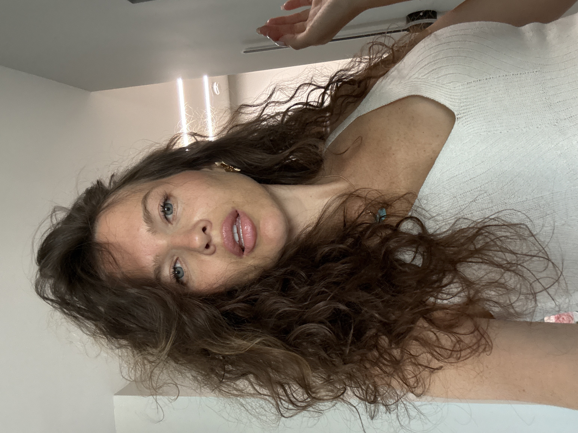 The image shows a close-up, self-taken portrait of a woman with long, curly brown hair and blue eyes. She is wearing a white, ribbed halter top. Her lips are slightly parted, and she has a faint smile. Her right hand is raised, with fingers extended, to the right side of the frame. The background appears to be an indoor setting, possibly a room with white walls and overhead lighting. There is no visible indication of the city of Khimki, Russia, in the image.