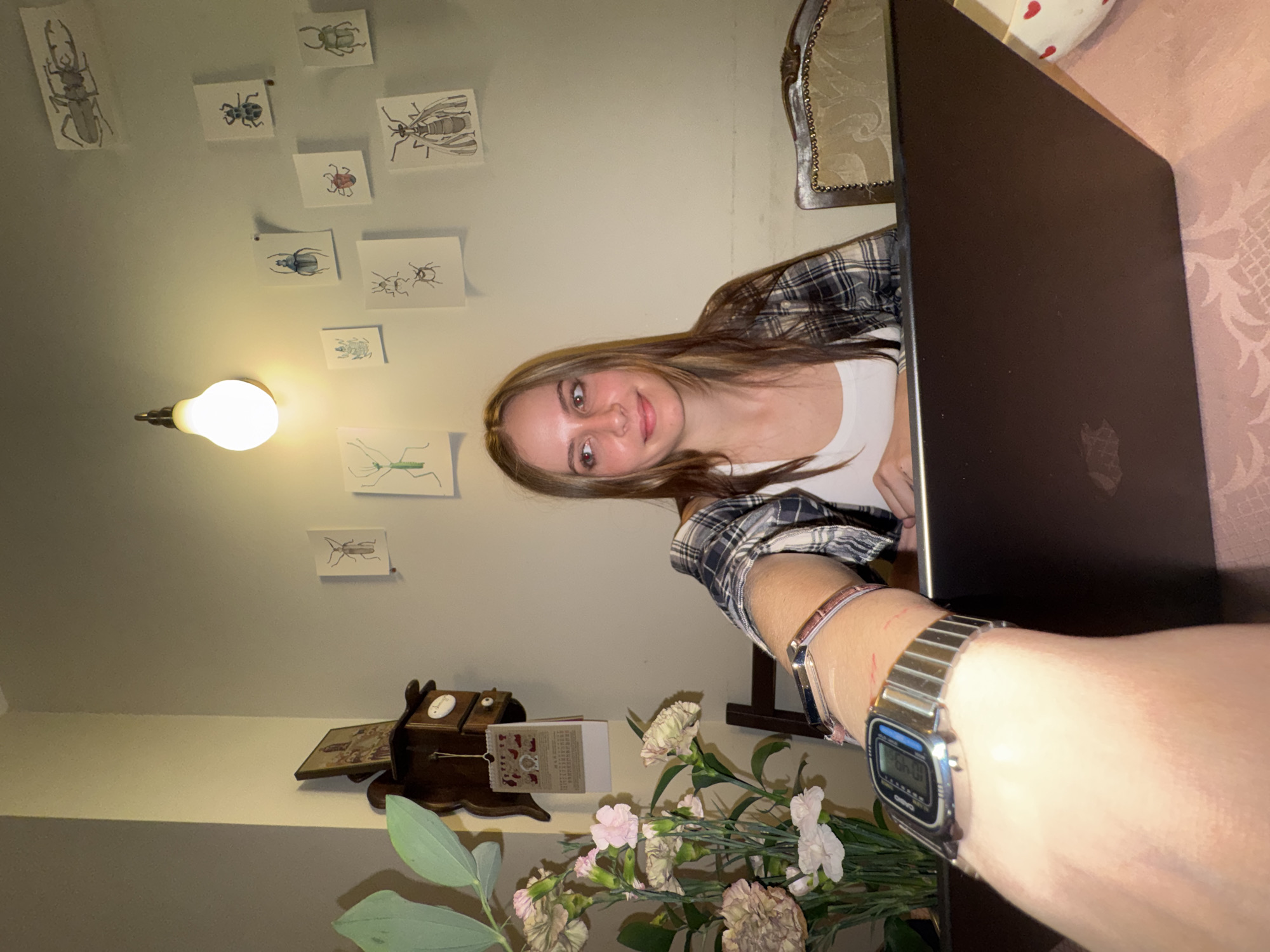 The image shows a young woman, likely in her late teens or early twenties, with long brown hair, sitting at a table indoors. She is wearing a plaid flannel shirt over a white top and is looking directly at the camera with a gentle smile. Her right arm, extending into the foreground, reveals two watches on her wrist: a silver digital Casio watch displaying "10:40" and a more delicate analog watch. An open, dark-colored laptop, possibly a MacBook with its distinct logo, rests on the table in front of her. The table is covered with a patterned tablecloth, and a white cushion with red heart patterns is visible behind the laptop.
To the woman's left, a light-colored wall is decorated with multiple small illustrations of various insects, including beetles, a mantis, and a wasp, illuminated by a single, exposed light bulb fixture. A classical-style wooden chair with ornate upholstery is partially visible behind her, to her right. In the foreground, to her right, a bouquet of light-colored flowers, possibly carnations, with green foliage, adds to the decor. Nearby, a dark wooden decorative item, possibly a small clock or box, sits on a surface beside a calendar with some illegible text. The scene suggests a relaxed indoor setting, potentially an evening in Moscow, Russia. The woman's expression is calm and friendly.