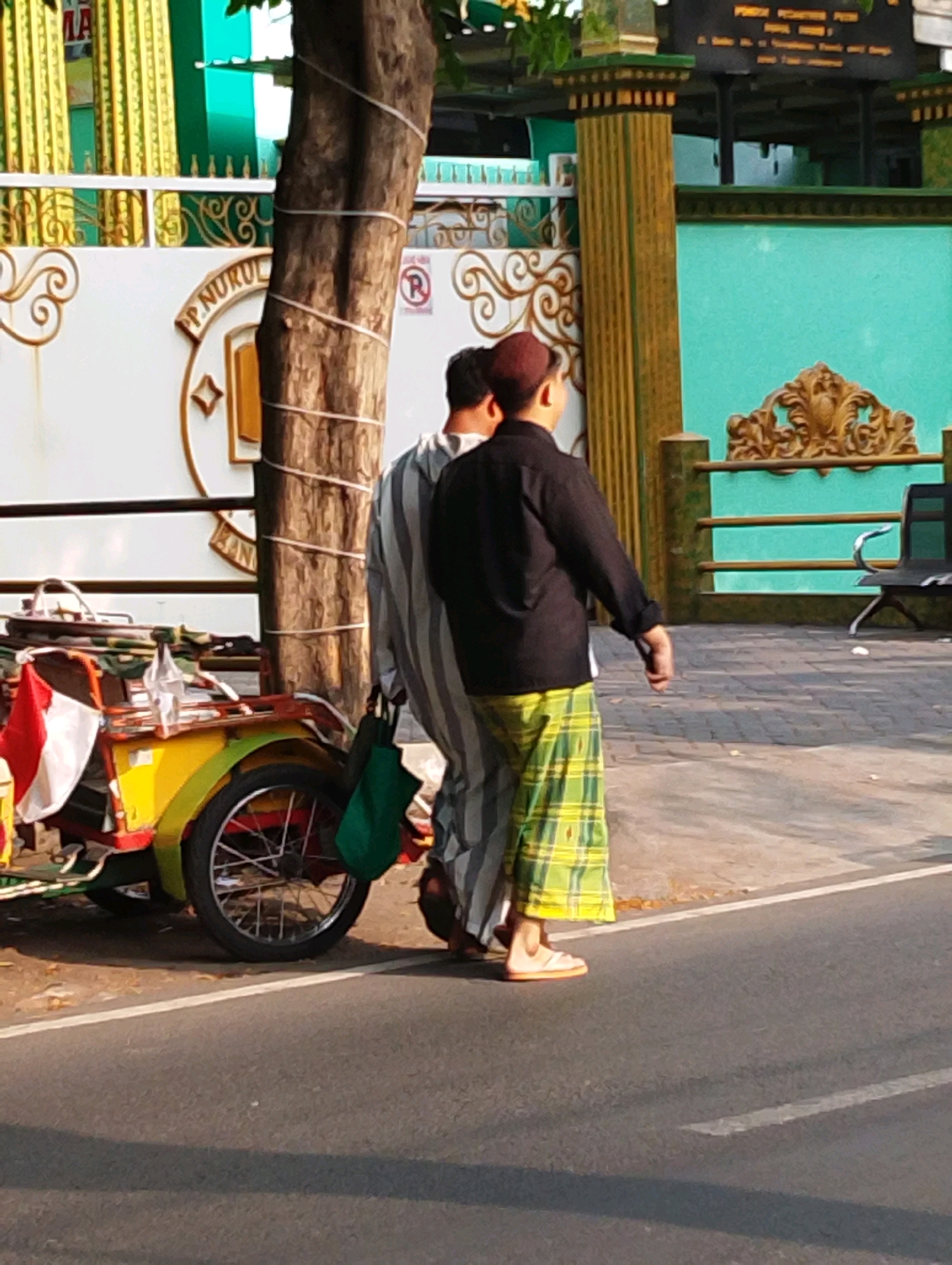Two individuals are standing next to a three-wheeled cart on a paved road. The individual on the left wears a striped garment and a maroon cap. The individual on the right wears a black long-sleeved shirt and a green plaid sarong. A green bag hangs from the left individual's shoulder. The cart is colorful, with yellow, red, and green accents, and has a red, white, and green flag attached to its front. Behind them, a wall with ornate gold trim separates the road from a building complex featuring teal and gold elements. The scene appears to be set in the city of Bangil, Indonesia, given the visible signage which includes "PP NURUL". The overall lighting suggests it is daytime, likely late afternoon given the shadows.