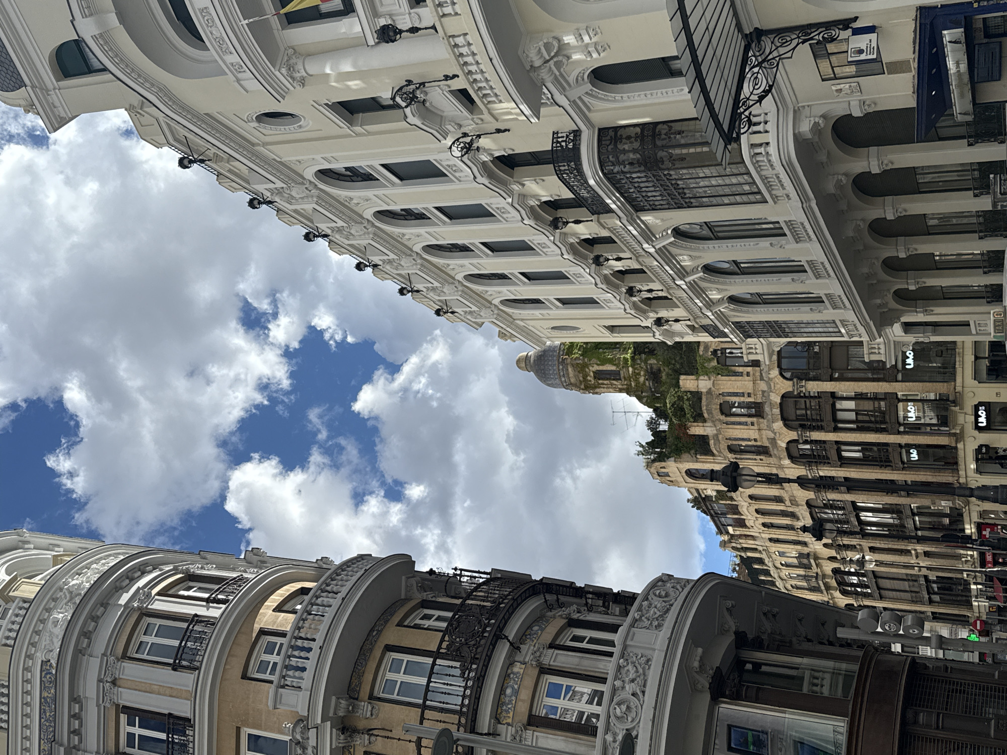 The image is taken from a low angle, showcasing buildings on the Gran Vía in Madrid, Spain, against a backdrop of a partly cloudy sky. The architecture is characterized by ornate facades with detailed moldings, arched windows, and decorative balconies. The buildings have varying tones, from white to beige and brown, reflecting different periods or styles. Streetlights and signage are visible, indicating a vibrant urban environment. The composition of the image gives a sense of grandeur and historical character to the street scene.