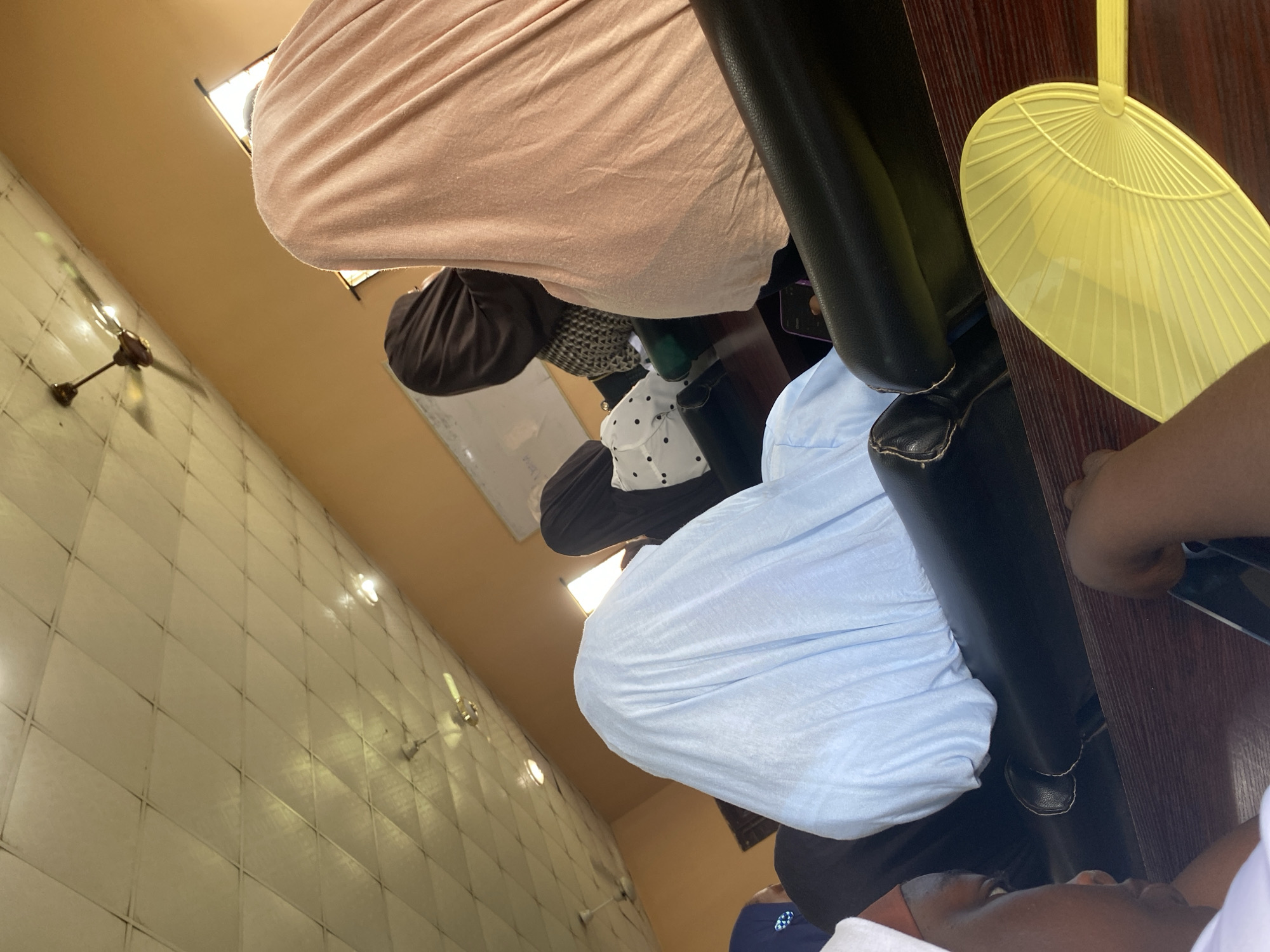 In an indoor setting in Sabo, Nigeria, a view angled upwards captures several seated individuals. One person wears a light pink garment. Another wears a light blue shirt, while a third is dressed in a black and white patterned outfit with a black hijab.
A yellow hand fan rests on a dark wooden table near a black leather chair. The wall features tiles. Overhead, a ceiling fan is visible.
