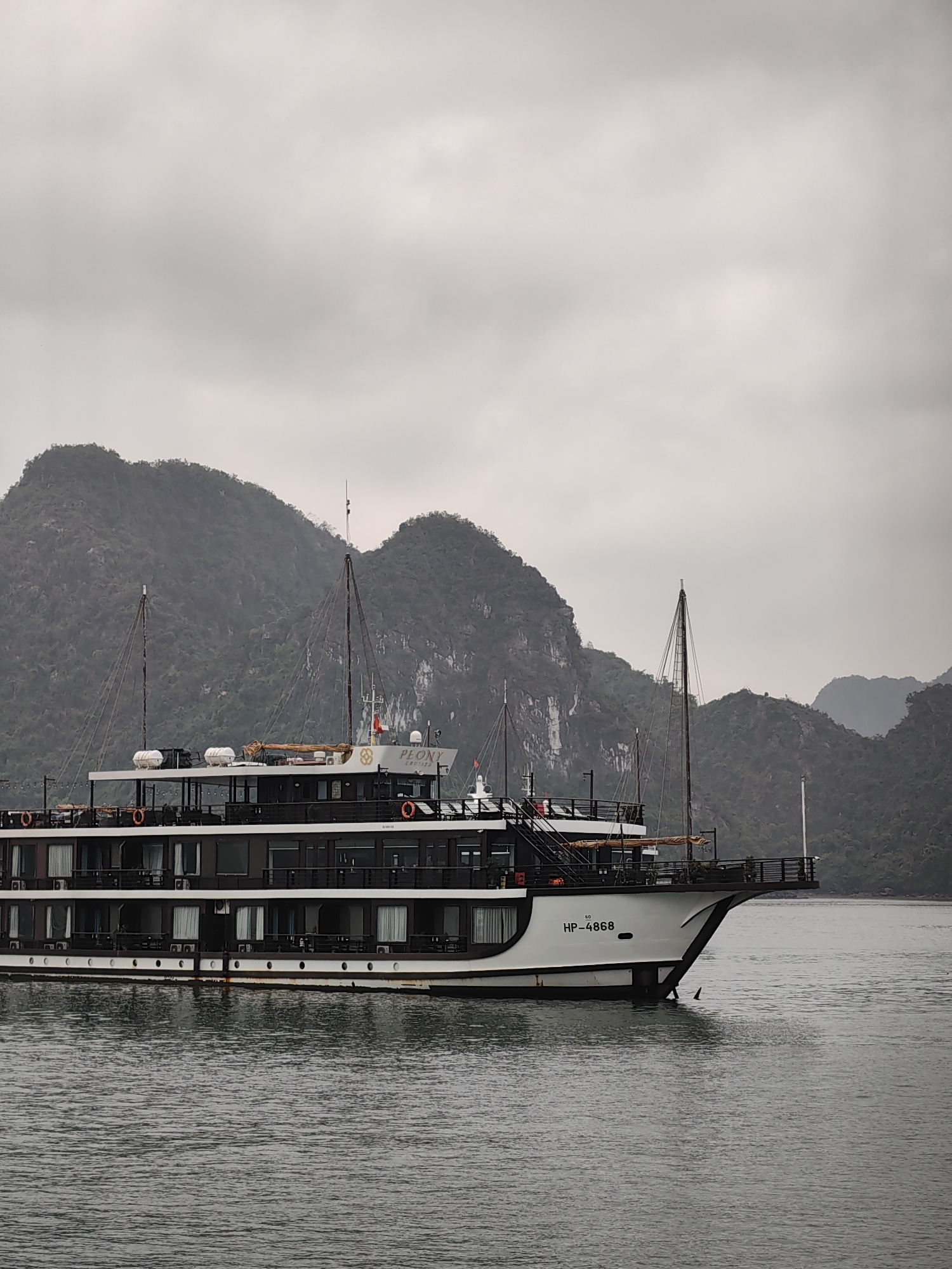 The image depicts a multi-deck cruise ship, possibly named "Peony Cruise" based on a partial view of the name on the ship's deck, sailing on the waters of Ha Long Bay in Hải Phòng, Vietnam. The ship is painted in black and white, with visible decks, windows, and life-saving equipment. The ship's hull displays the registration number "HP-4868." In the background, there are limestone karsts characteristic of the Ha Long Bay landscape, shrouded in mist. The sky is overcast, indicating a cloudy day. The water is calm and gray. The overall impression is a landscape view of Ha Long Bay with a focus on a cruise ship navigating its waters.
