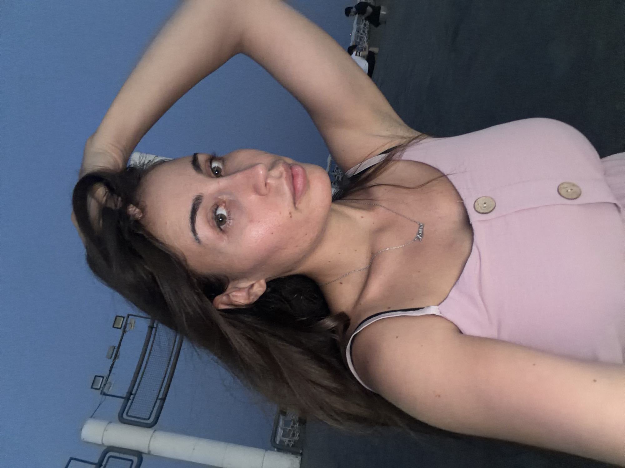 A selfie of a woman with long, dark brown hair and brown eyes is taken outdoors at dusk. She is wearing a pink spaghetti-strap dress with two light brown buttons down the front. A silver necklace with a script name is visible. She is holding her right hand to her hair. In the background, a white cylindrical structure and lighting fixtures are visible. Further in the background, two individuals are seated on what appears to be beach chairs, with another person standing in the distance. The sky is a deep blue, indicating twilight. The scene is likely a public outdoor area near the coast, possibly in Larnaca, Cyprus, given the characteristic coastal infrastructure and lighting found in such locations.