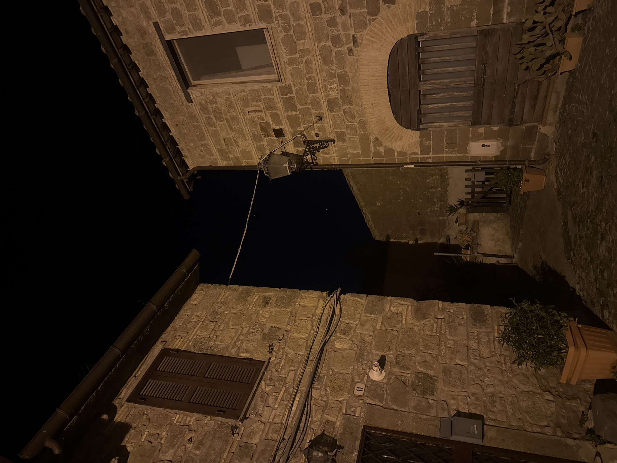 The image shows a narrow street in Castelnuovo di Porto, Italy, at night. The perspective is angled upward, showing the facades of two buildings constructed of rough-hewn stones, facing each other across a narrow space. Above, the sky is dark. The buildings feature windows with closed shutters and one arched window. Streetlights illuminate the stone walls. Several potted plants sit at ground level or on walls. Electrical wires span the gap between buildings.