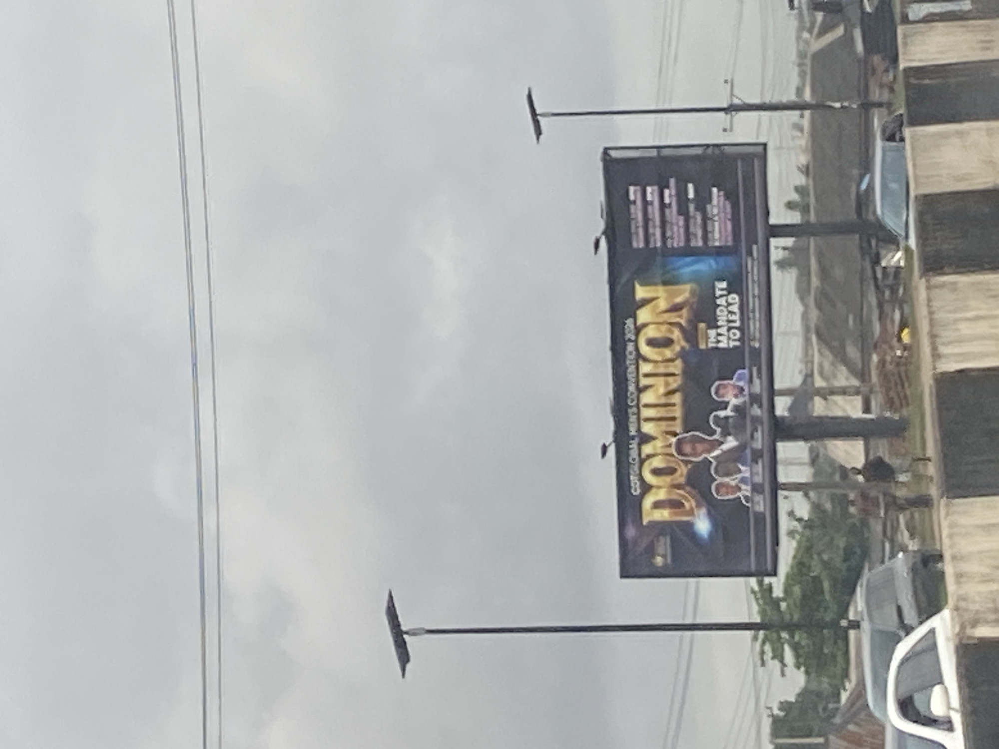 A wide shot captures a billboard in the city of Ikorodu, Nigeria, advertising "DOMINION" and "THE MANDATE TO LEAD" for the "CGTGLOBAL MEN'S CONVENTION 2026." The billboard features images of several men in formal attire. The scene is overcast, with visible power lines and streetlights. In the foreground, a concrete barrier lines a road where several cars are visible, including a white car with its driver's side door open. Two individuals stand near a construction site behind the barrier. In the background, buildings and trees are partially obscured by haze and distance.