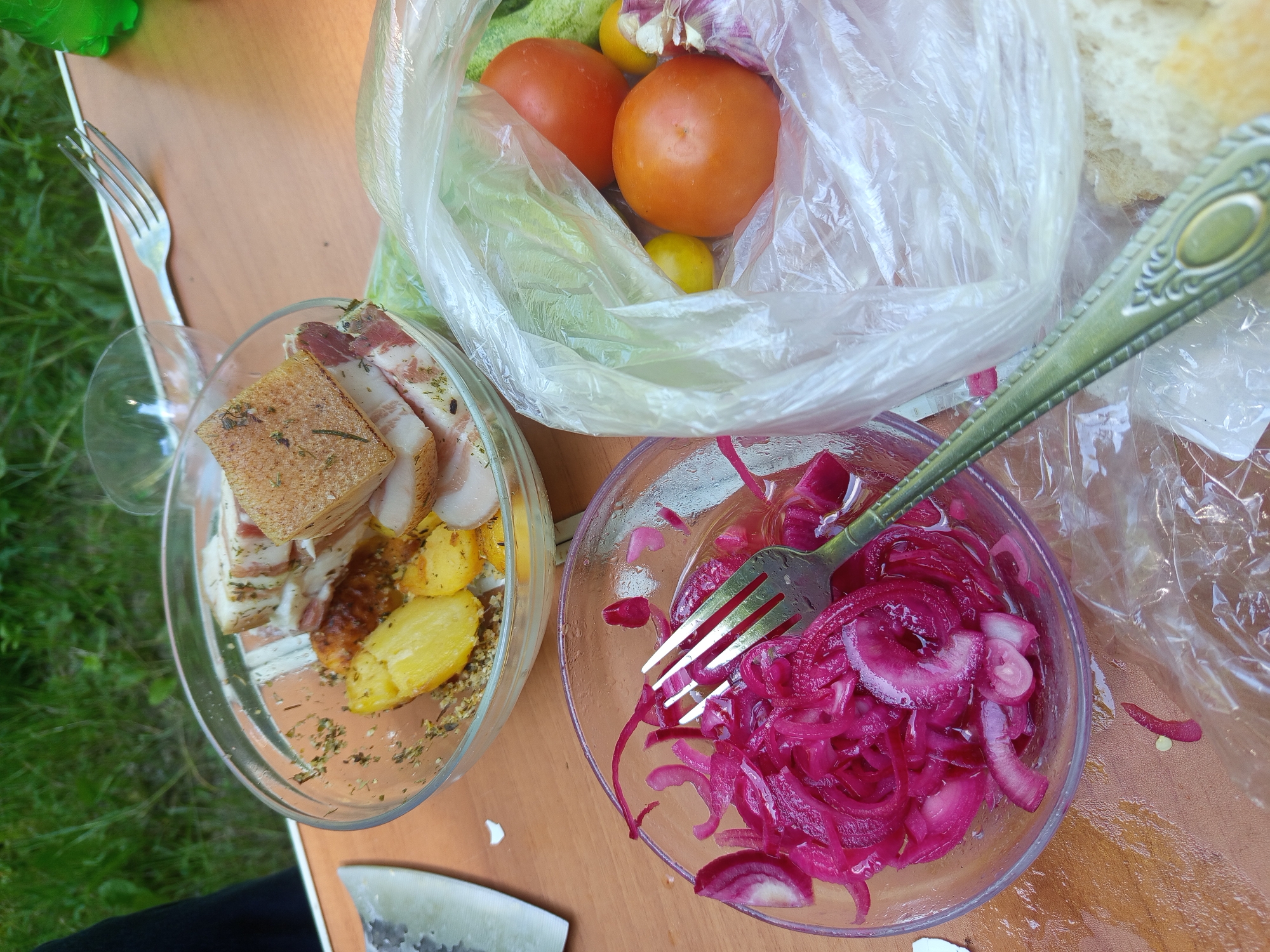 The image displays a rustic outdoor setting, likely a picnic or meal outdoors, with food laid out on a wooden surface. In the foreground, a clear bowl contains sliced red onions that appear to be pickled or marinated, with a fork resting inside. To the right of this, a transparent plastic bag holds various vegetables, including two ripe tomatoes, what appear to be cucumbers, a head of garlic, and a couple of small yellow fruits, possibly lemons or small tomatoes. Behind these items, slices of bread are visible. In the background, another clear bowl contains roasted potatoes and slices of what looks like cured meat or bacon, seasoned with herbs. A knife is positioned to the left of the bowls, and a fork is visible on the table behind the first bowl. The environment suggests a natural setting with greenery visible in the upper background, and the wooden surface indicates a table. The overall impression is one of a casual, freshly prepared meal. Given the context provided of the city of Giaginskaya, Russia, this scene likely depicts a common outdoor dining practice in a rural or suburban area of the region.