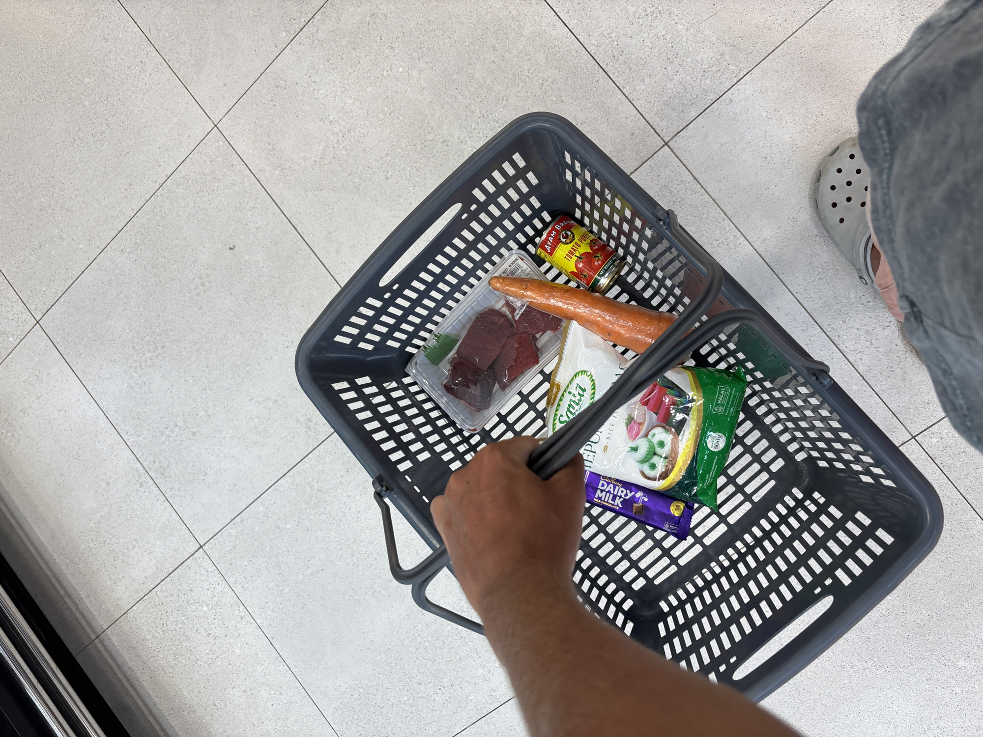 An overhead shot captures a person shopping at what appears to be a grocery store or supermarket in Kuta Selatan, Indonesia. The primary subjects are a person's right hand gripping the handle of a grey plastic shopping basket, and their lower right leg and foot, clad in a light grey Croc shoe, visible on the right side of the frame.
Inside the shopping basket are several grocery items. These include a red can of "AYAM BRAND TOMATO PUREE," a clear plastic tray containing raw red meat (likely beef), a single large carrot, a white bag with green lettering indicating "Sania RICE FLOUR," a green packaged item marked "HALAL" and "500G," and a purple wrapper for a "Cadbury DAIRY MILK" chocolate bar.
The floor is a light grey tile with dark grout lines, characteristic of a retail environment. The activity taking place is grocery shopping, with the person having already selected a few items for purchase. The presence of the "HALAL" certification on one package and the "AYAM BRAND" tomato puree are consistent with the provided location context of Indonesia. The time of day and weather are not discernible, and no specific emotions are conveyed.