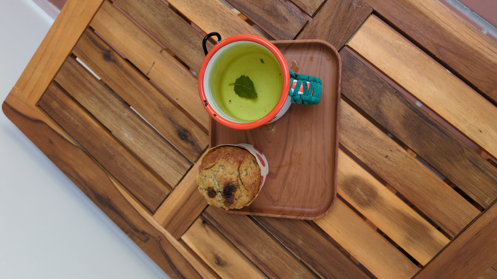 A high-angle shot depicts a cup of tea and a muffin presented on a wooden table with a slatted surface. The tea is contained within a white enamel mug, rimmed with orange and featuring a paracord-wrapped handle. A single mint leaf floats on the tea's surface. The muffin, nestled in a paper cup adorned with a strawberry pattern, rests beside the cup on a wooden tray. The table's wooden slats create a textured background, suggesting an outdoor setting. This likely captures a quiet moment in Da Lat, Vietnam, where such scenes are common due to the city's climate and cafe culture.