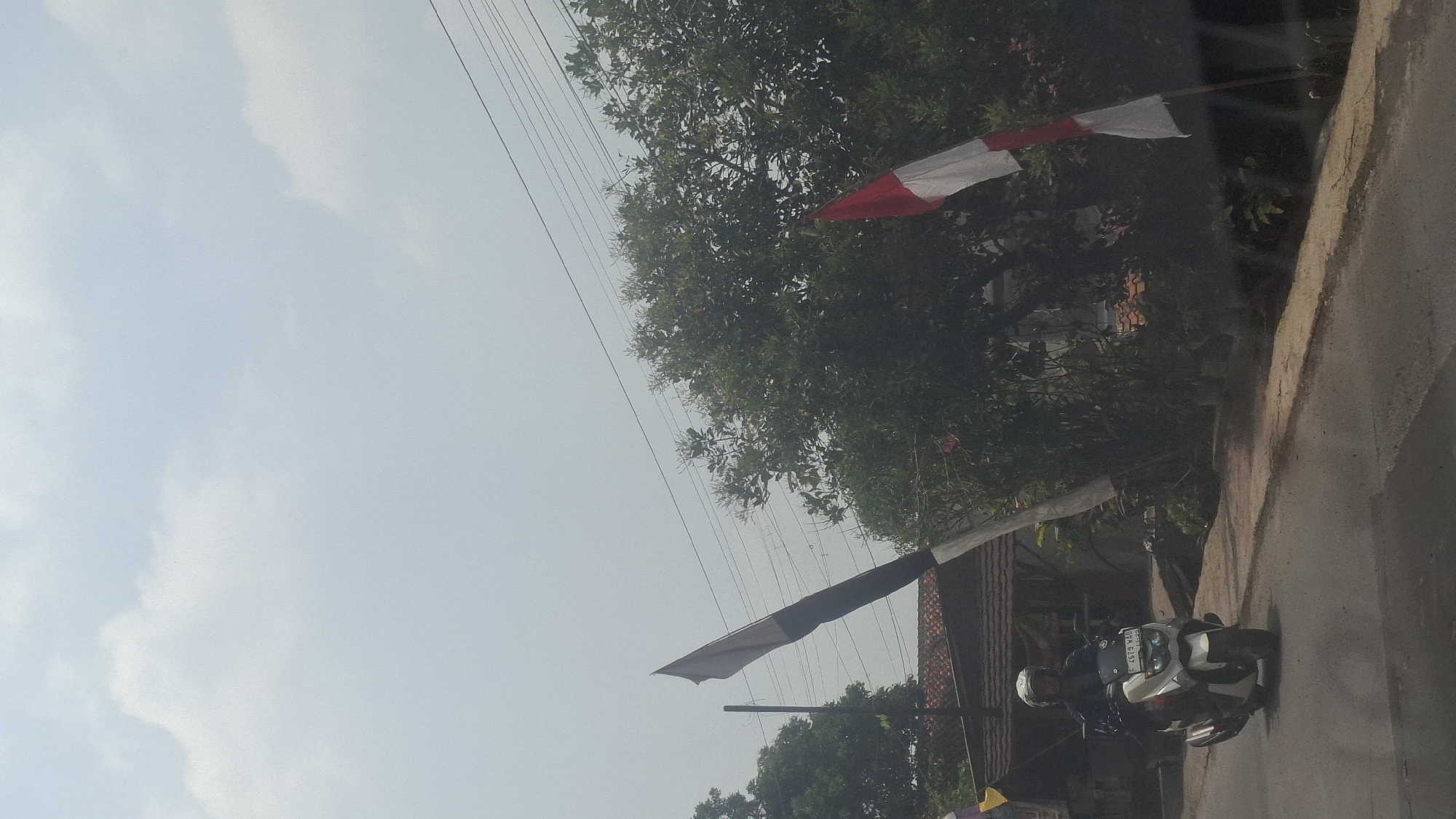 In Sirnagalih, Indonesia, a person wearing a helmet is riding a white scooter on a paved road. The scooter has a license plate reading "F 4679 WAD". To the right of the road, there are trees and foliage. Two flags are visible: one is a long, striped flag, dark at the bottom and lighter at the top, attached to a pole. Further to the right, a red and white flag is mounted on a pole, partially obscured by trees. In the background, there are buildings with tiled roofs and utility wires are strung overhead. The sky above is overcast with white clouds.