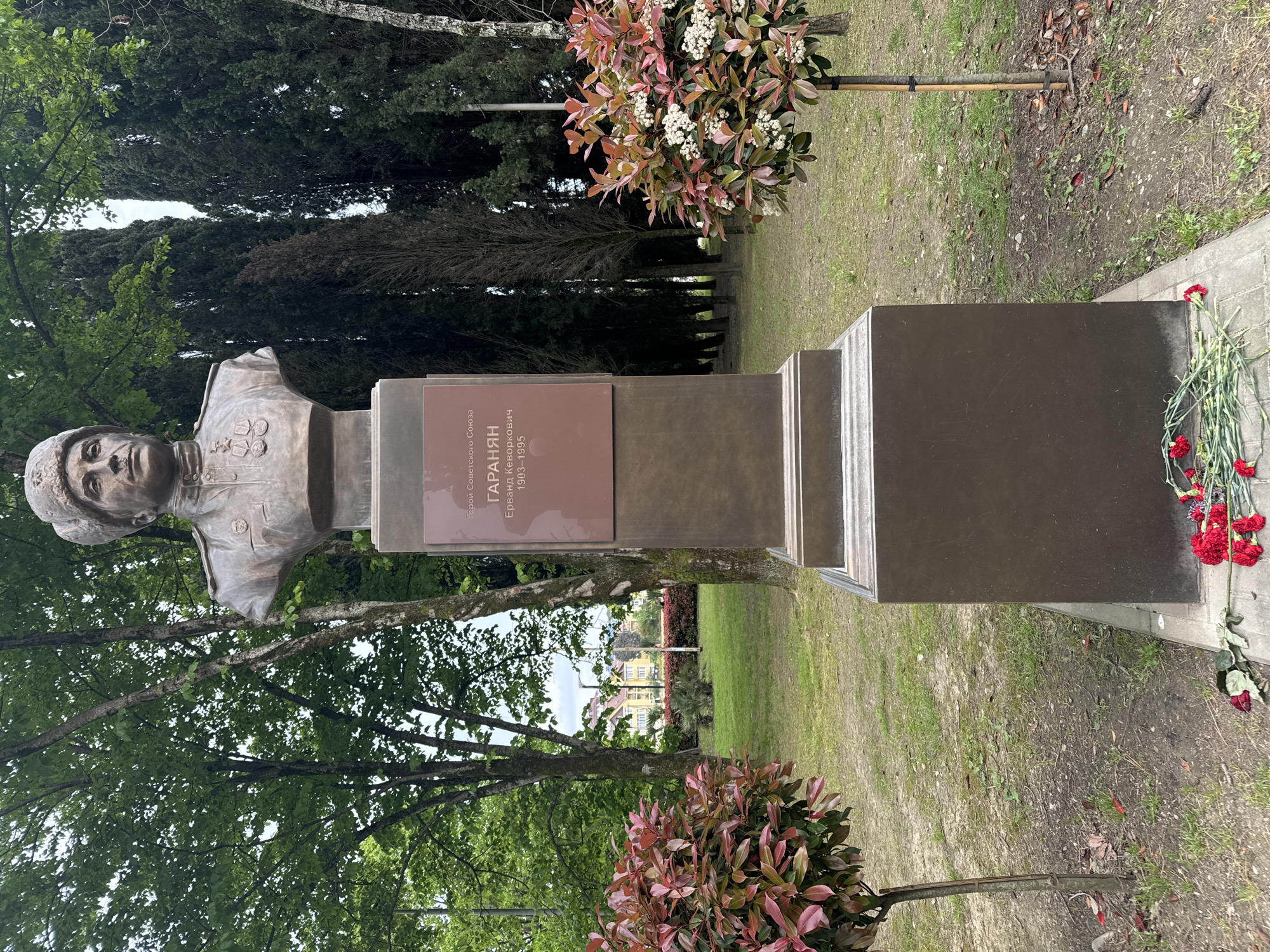 A bronze bust of a man in military uniform is mounted on a tiered pedestal. A plaque on the pedestal identifies the subject as Герой Советского Союза Гараян, Ерманд Кеворкович (Hero of the Soviet Union Garayan, Ermand Kevorkovich) with the dates 1903-1995. The bust faces left. Red carnations lie at the base of the pedestal. Shrubs with red and green leaves flank the base of the monument. Trees and green grass are visible in the background. The photo appears to have been taken in daylight at Sochi, Russia.