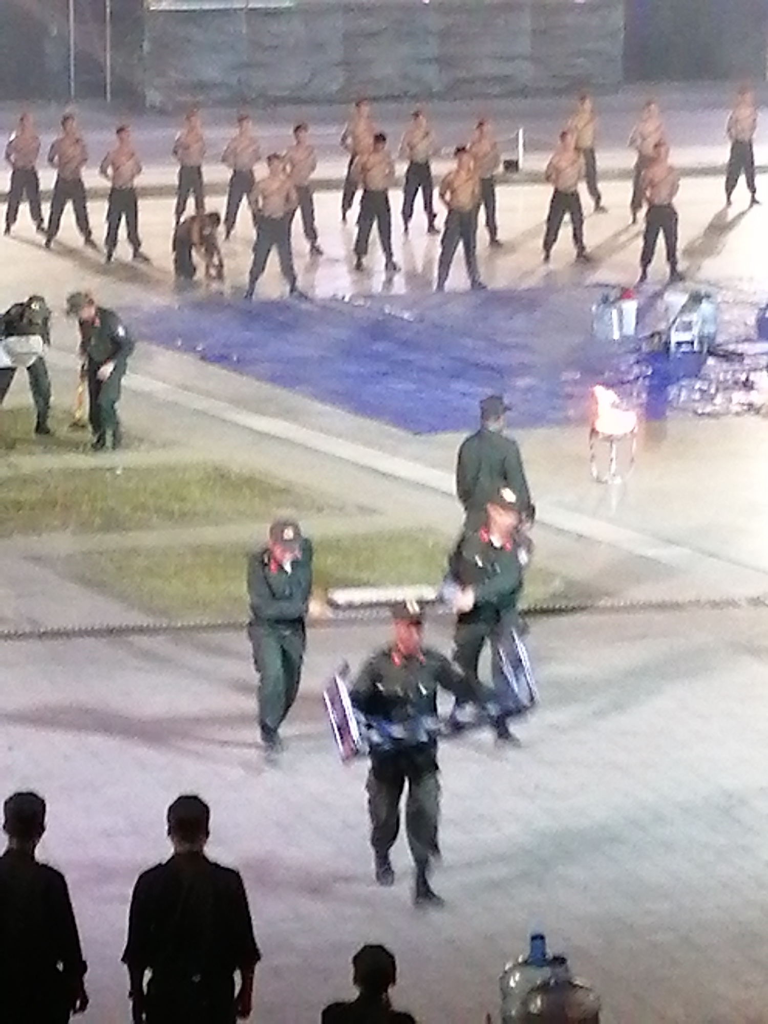 The image depicts a group of people, possibly soldiers, engaged in an outdoor activity. Several individuals are shirtless, wearing dark pants, and appear to be performing some synchronized movements. Some are holding shields, suggesting a drill or performance. In the background, a group of similarly dressed individuals are also present. A large blue tarp or mat is laid out on the ground, and a flaming object is visible in the distance, possibly a torch or a ceremonial fire. The lighting suggests it is either dusk or nighttime. The scene appears to be an open area, possibly a training ground or an arena. There are no visible text elements in the image.
