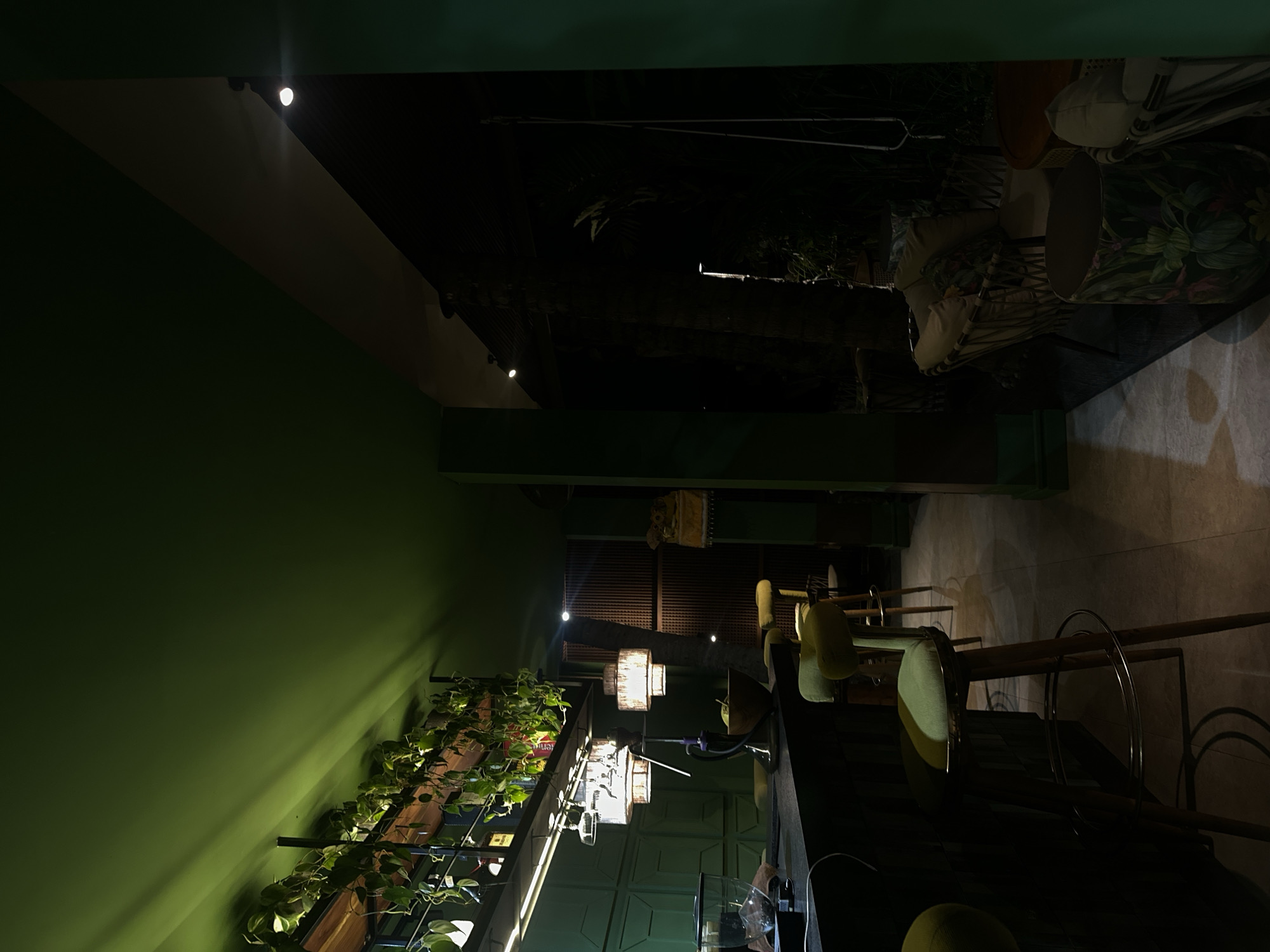 The image depicts a dimly lit bar or restaurant interior, possibly in Ubud District, Indonesia. The walls are painted a muted green, with a section featuring a vertical garden with trailing plants. Hanging lights illuminate the bar area, where tall bar stools are positioned. The setting is intimate and modern, with a focus on greenery. Beyond the bar, the space opens up into a darker area with chairs and what appears to be a partially visible outdoor space or another section of the restaurant.