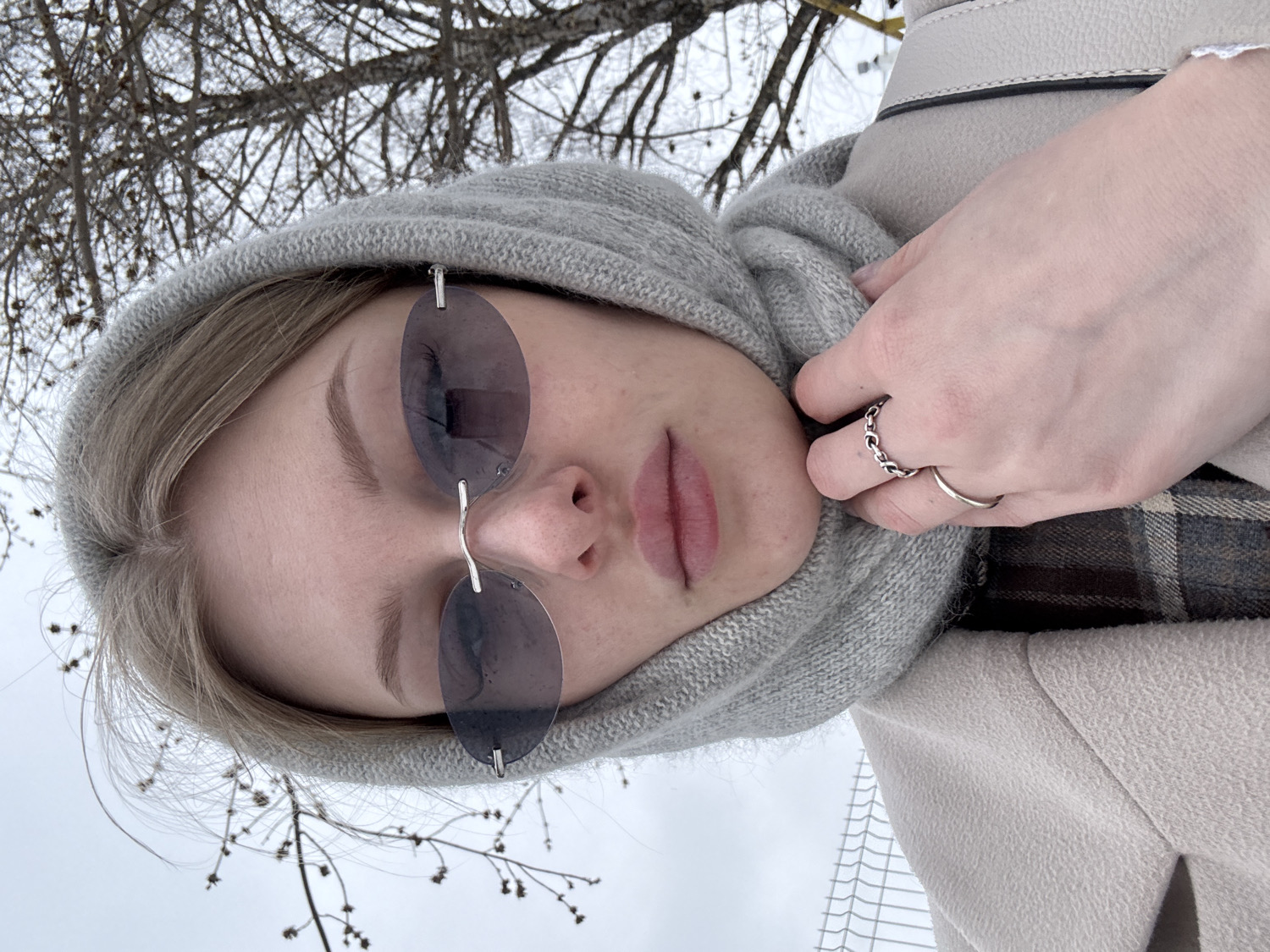 A young woman is the central subject of this tilted outdoor portrait, likely taken as a selfie. She wears a gray knitted hooded scarf that frames her face and light-colored hair. Her eyes are covered by rimless oval sunglasses with dark lenses. Her lips are slightly parted, and her expression appears neutral. Her left hand rests near her chin, adorned with two silver rings: one a decorative chain-link design and the other a simple band. She is dressed in a light beige coat, and a brown and gray plaid pattern, possibly part of an undergarment or scarf, is visible near her shoulder.
The scene is set outdoors in a winter landscape, specifically in Perm, Russia, which is evident from the bare, dark tree branches creating a delicate, intricate pattern against an overcast, bright white sky in the background. The overall color palette is muted, reflecting a cold, subdued atmosphere. The weather is likely cold and overcast. No text is visible in the image.