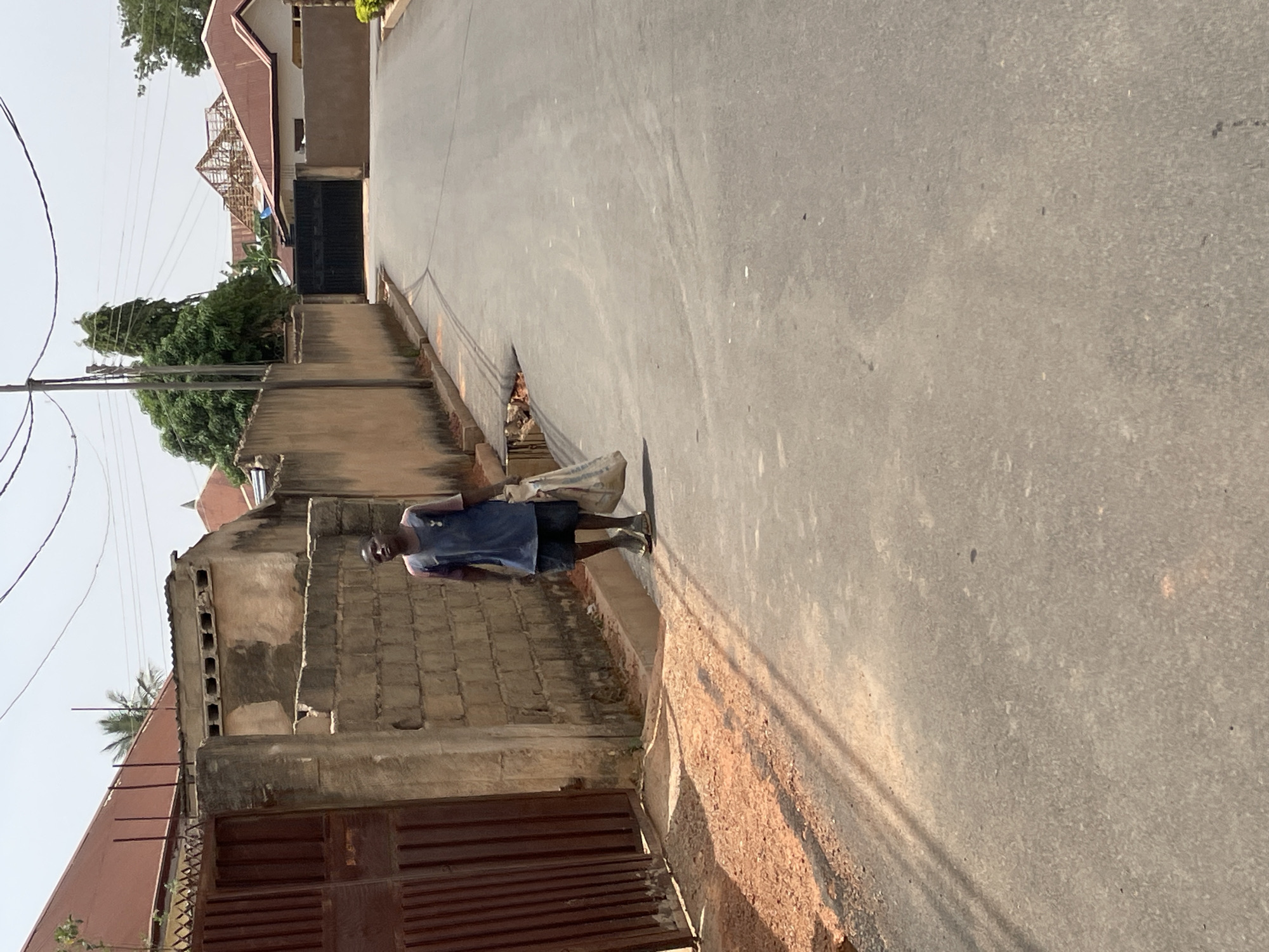 A man is walking down a narrow paved road in Jos, Nigeria. The road is bordered by concrete and brick walls. The man is wearing a dark shirt and shorts, and walking away from the camera. A building with a brown roof and metal gate is seen on the left side of the road. Overhead power lines are visible. The weather appears sunny.
