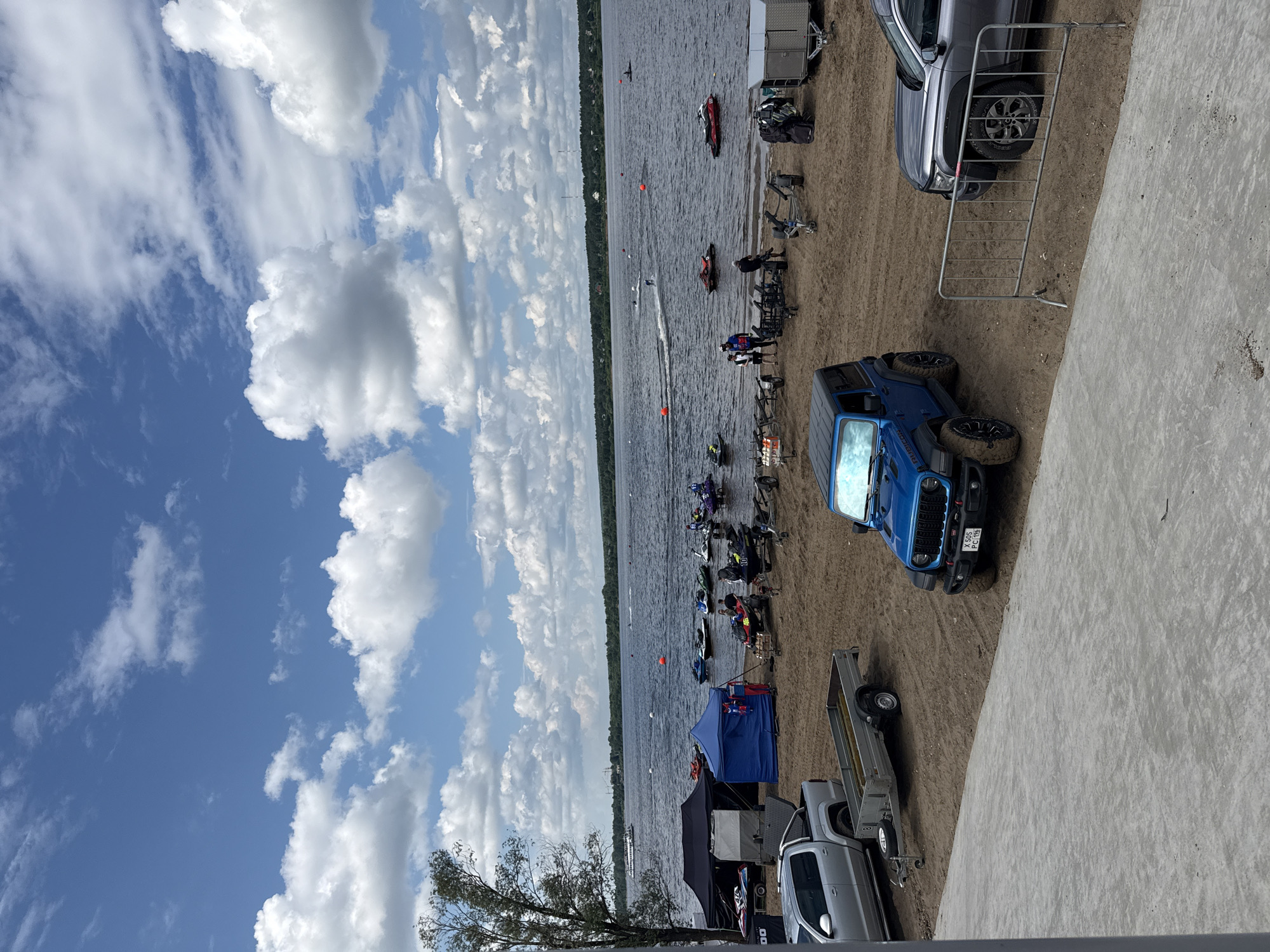 The image depicts a sunny day at a waterfront location, likely in Perm, Russia, with a wide river or bay in the background and a sandy shore in the foreground. The water is active with several jet skis, some in motion and others docked or on trailers. A variety of tents are set up on the sand, possibly for an event or race.
In the foreground, three vehicles are visible: a silver pickup truck with its tailgate down, a blue Jeep Wrangler with a license plate that reads "X 505 PC 196", and a dark gray SUV. A metal barrier separates the sandy area from a paved walkway in the immediate foreground.
Several people are scattered around the scene, some near the jet skis, others in the vicinity of the tents, and some appear to be interacting with the vehicles. The sky is blue with scattered white clouds, and a large boat is visible in the distance on the water, along with a treeline and buildings on the far bank. The overall atmosphere suggests an outdoor recreational event.
