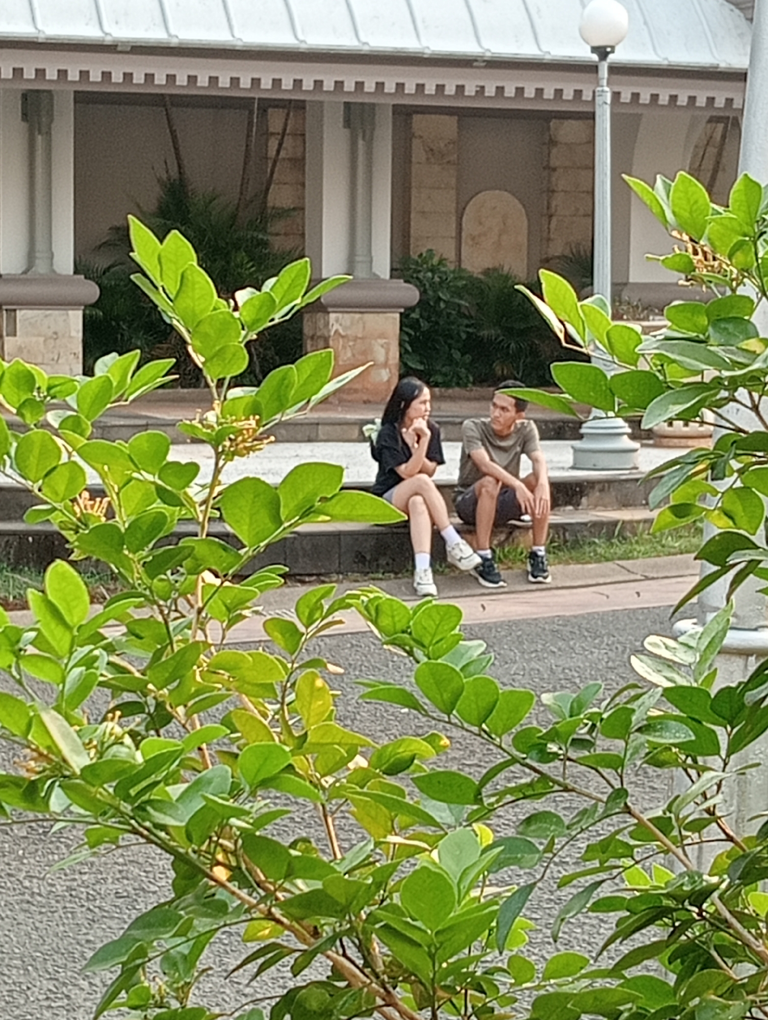 In an outdoor setting, two individuals are seated on the steps of a stone structure. A woman, with long dark hair, wears a black t-shirt and light-colored shorts. She is seated with her legs crossed and her hands resting near her chin. Beside her, a man with short dark hair is wearing a brown t-shirt and dark shorts. He is sitting with his legs extended and his arms resting on his knees. Both individuals appear to be engaged in conversation, facing each other. The background features a building with columns and a white dome-like roof, surrounded by greenery and foliage. A lamppost is visible to the right of the building. The scene suggests a public park or a similar outdoor public space.