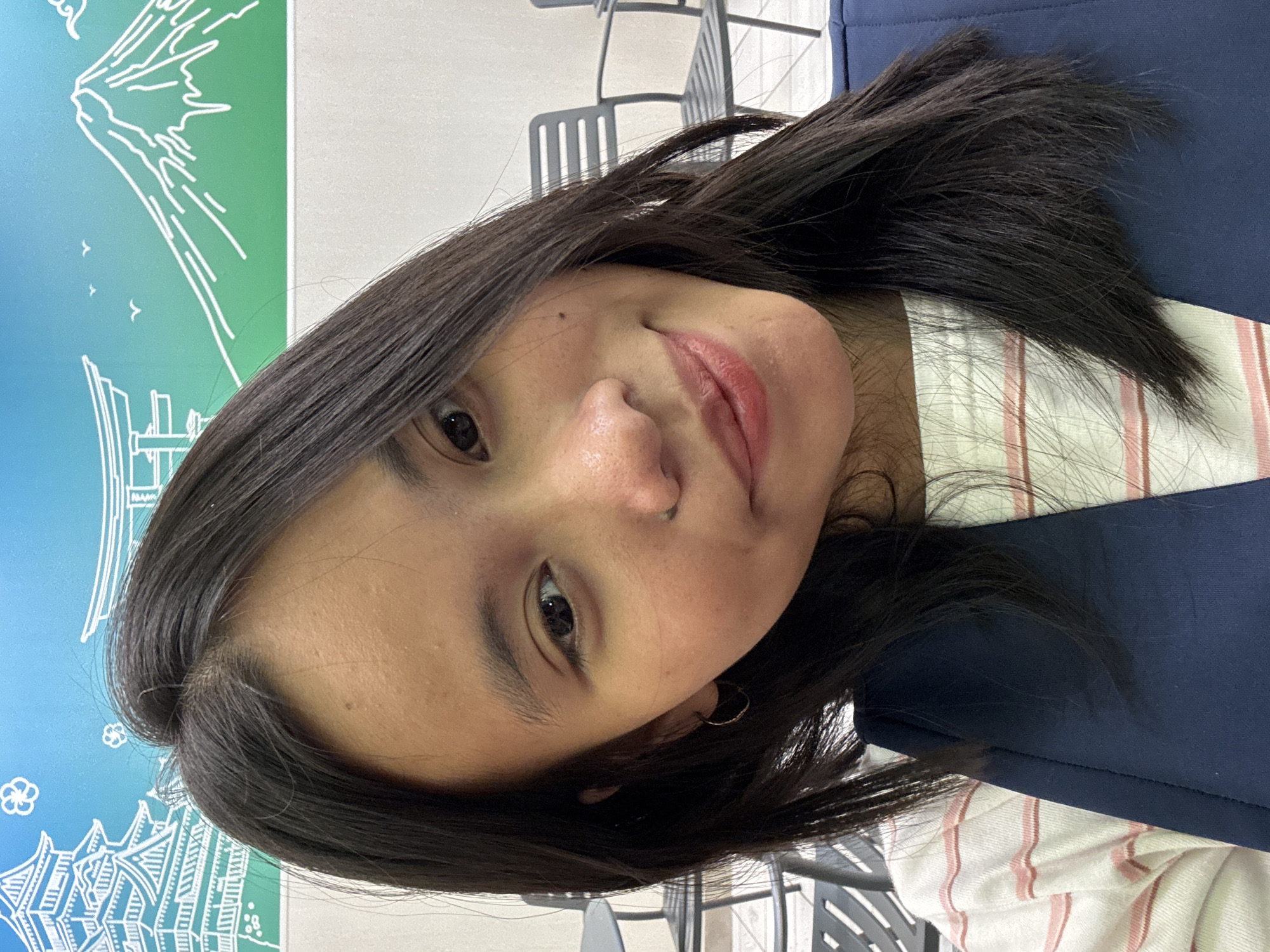 A medium shot captures a young woman with dark, straight hair from a selfie angle. She is positioned in the foreground, with her head tilted slightly to her right. She has brown eyes, a small nose with a subtle nose ring, and is smiling gently, her lips appearing to have a light tint. A small mole is visible on her left cheek. She is wearing a white t-shirt with thin pink stripes and a dark blue vest or jacket over it.
The background features a wall mural depicting a stylized Japanese landscape, with a blue sky at the top. On the left side of the mural, a white outline of a multi-tiered pagoda-like structure is visible, with small white floral motifs. On the right, a white outline of a mountain, resembling Mount Fuji, is depicted against a green background.
In the middle ground, out of focus, are several dark gray slatted chairs, suggesting an indoor seating area. The lighting is even, and the overall impression is of a casual, indoor setting.
The image is not taken in East Jakarta, Indonesia. It depicts a backdrop with Japanese cultural motifs.