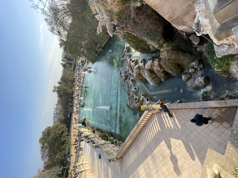 The image displays an elevated view of the Cascada Monumental (Monumental Waterfall) in Parc de la Ciutadella, Barcelona, Spain, under a clear blue sky.
The central feature is a large, turquoise-blue artificial pond in the midground, containing multiple operational fountains with water jets spraying upwards. This pond is bordered on the left by an ornate stone balustrade, featuring decorative urns on its posts. Beyond the pond, a wide park area is visible with numerous deciduous and palm trees, and dozens of people are scattered across pathways.
To the right of the main pond, water flows over sculpted rock formations and smaller waterfalls into a lower, narrower water channel. Integrated into the rockwork on the far right, a classical stone sculpture of a reclining figure, partially obscured, is visible.
In the foreground, a wide, light-colored tiled pathway extends diagonally from the bottom left. A person wearing a dark blue jacket and carrying a dark shoulder bag walks away from the viewer on this path. Further along the stone balustrade bordering this pathway, another individual in a dark jacket with a red hood is leaning over the railing, looking down towards the lower waterfalls.
The scene is brightly lit by natural sunlight, casting long shadows from the balustrade and individuals onto the pathway, indicating a position of the sun likely in the mid-morning or late afternoon.
