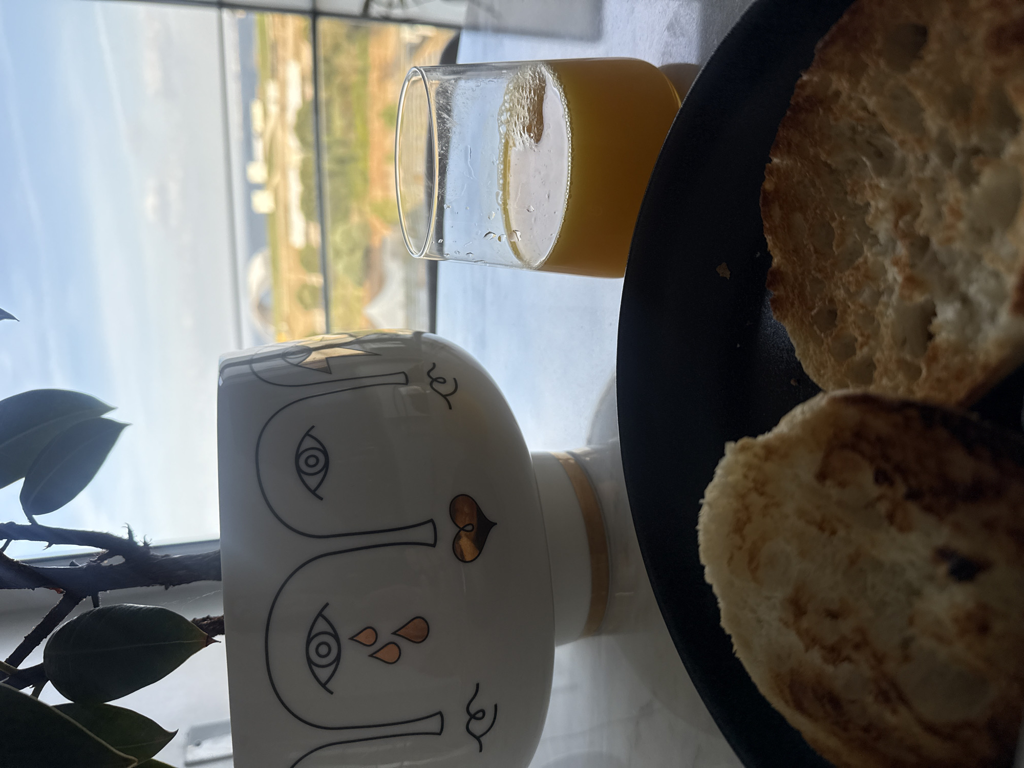 A photograph depicts a breakfast scene with a view visible through a window. The foreground features a white ceramic vase decorated with an abstract face design, a glass of orange juice, and a black plate with two pieces of toasted bread. A plant with dark green leaves is positioned beside the vase. The background view from the window shows a landscape with a mix of buildings and vegetation. The location is in the city of Loures, Portugal.