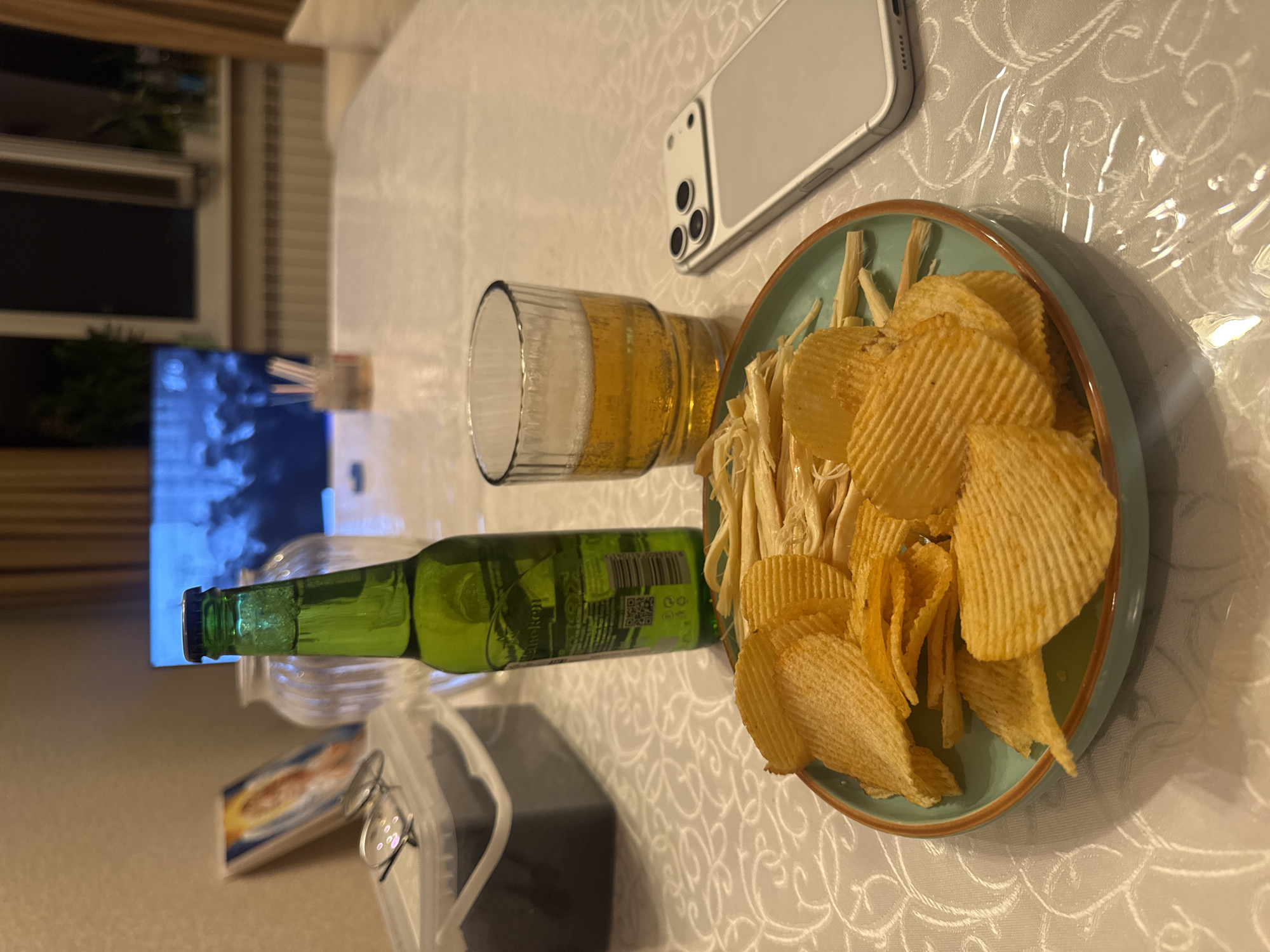 A close-up, eye-level shot shows a table with food and drink. In the foreground, a light blue plate holds a pile of ruffled potato chips and dried fish strips. To the right of the plate is a clear glass filled with beer and a white smartphone with three camera lenses. Behind the plate and glass stands a green bottle of Heineken beer. In the background, a clear glass vase, a small framed picture, and a pair of glasses sit on a white tablecloth. Further in the background, a television displays a dimly lit image. The scene appears to be indoors, possibly in a home environment. There are no people visible in the image. The lighting is dim, and the overall atmosphere suggests a casual setting. The location is specified as the city of Almaty, Kazakhstan.