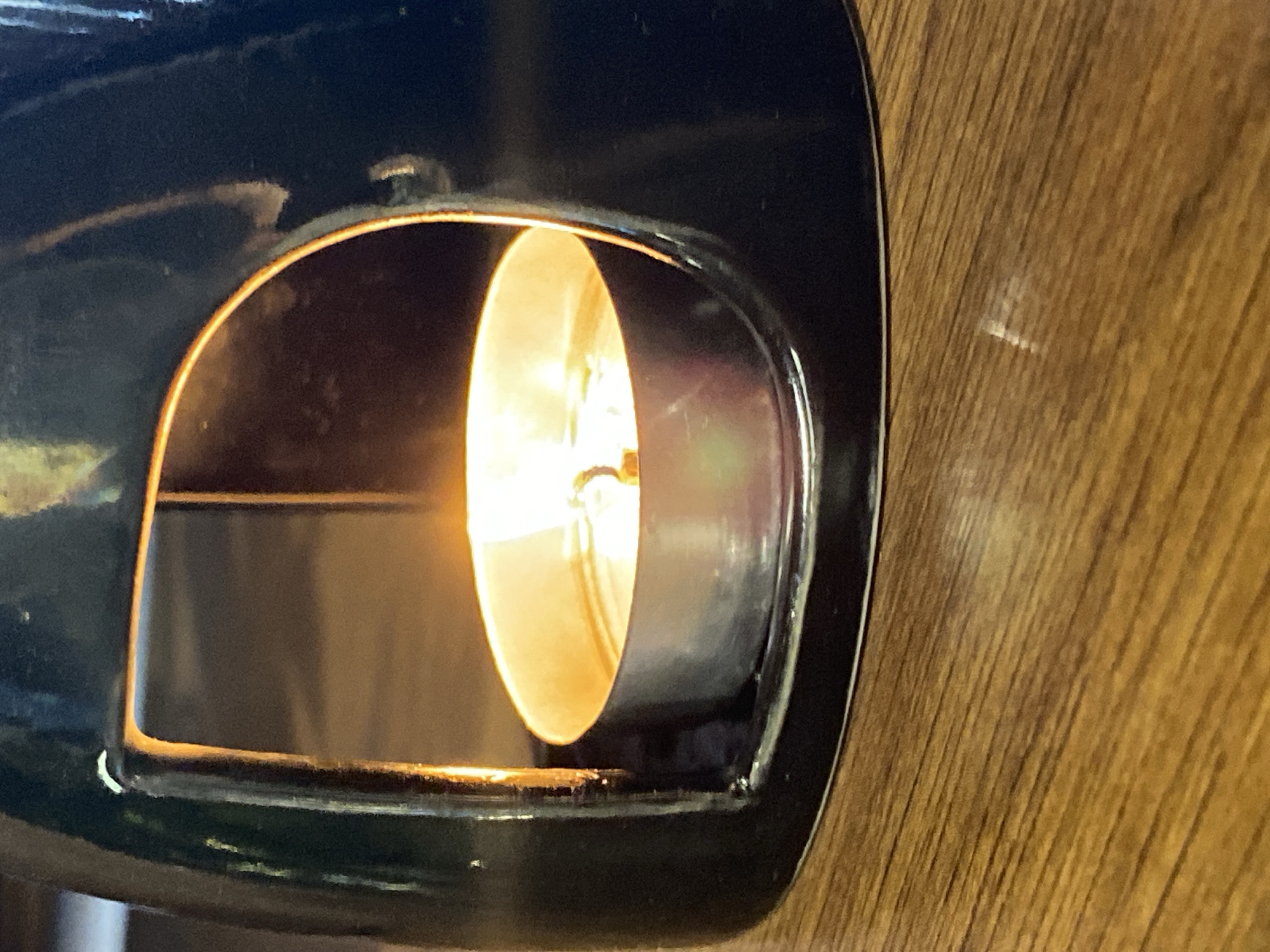 A dark, glossy, possibly ceramic or glazed candle holder, appearing very dark blue or black, sits on a polished wooden surface in the city of Votkinsk, Russia. The holder features a large, arched opening on its front, revealing a lit tealight candle positioned within its base. The tealight, contained in a silver-colored metal cup, emits a bright, warm orange-yellow flame from a central wick. Light from the flame illuminates the interior of the holder and reflects off its glossy exterior, particularly around the opening, and off the metallic rim of the candle cup. The wooden surface displays distinct linear grain patterns, running predominantly horizontally across the lower foreground, and reflects a faint, diffuse light source. The background is out of focus, exhibiting muted, warm brown tones. No people are visible.