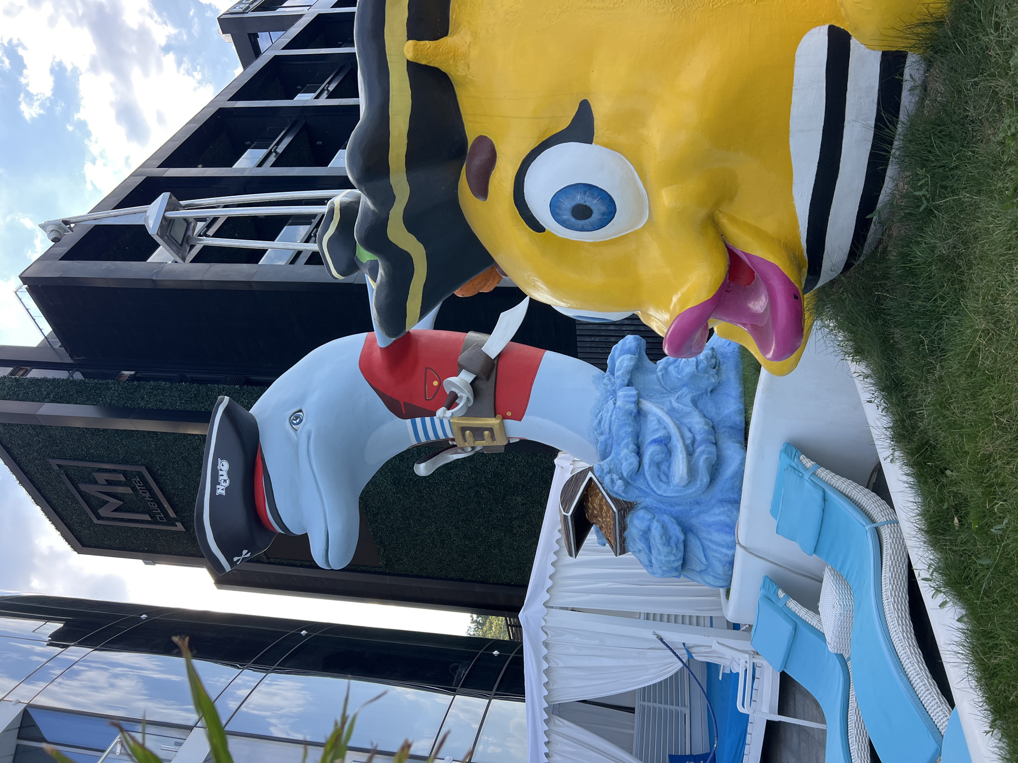 This is a medium shot of a sunny outdoor scene at the M1 Club Hotel in Odesa, Ukraine, with a focus on two large, colorful, cartoonish sculptures. In the foreground to the right is a bright yellow fish sculpture with wide blue eyes, pink lips, and a black and white striped body. To the left of the fish is a light blue dolphin sculpture dressed as a pirate, complete with a pirate hat and a red coat. The dolphin is perched on a blue, wave-like sculpture that has a treasure chest overflowing with gold coins at its base.
Behind the sculptures, a modern, dark-colored building with multiple balconies is visible. To the left of that, a tall building with a reflective glass exterior stands. In the mid-ground, a white tented cabana with white curtains is set up, with blue lounge chairs visible inside. In front of the cabana are two white wicker lounge chairs with bright blue cushions and pillows. The ground is covered in green grass. The sky is partly cloudy.