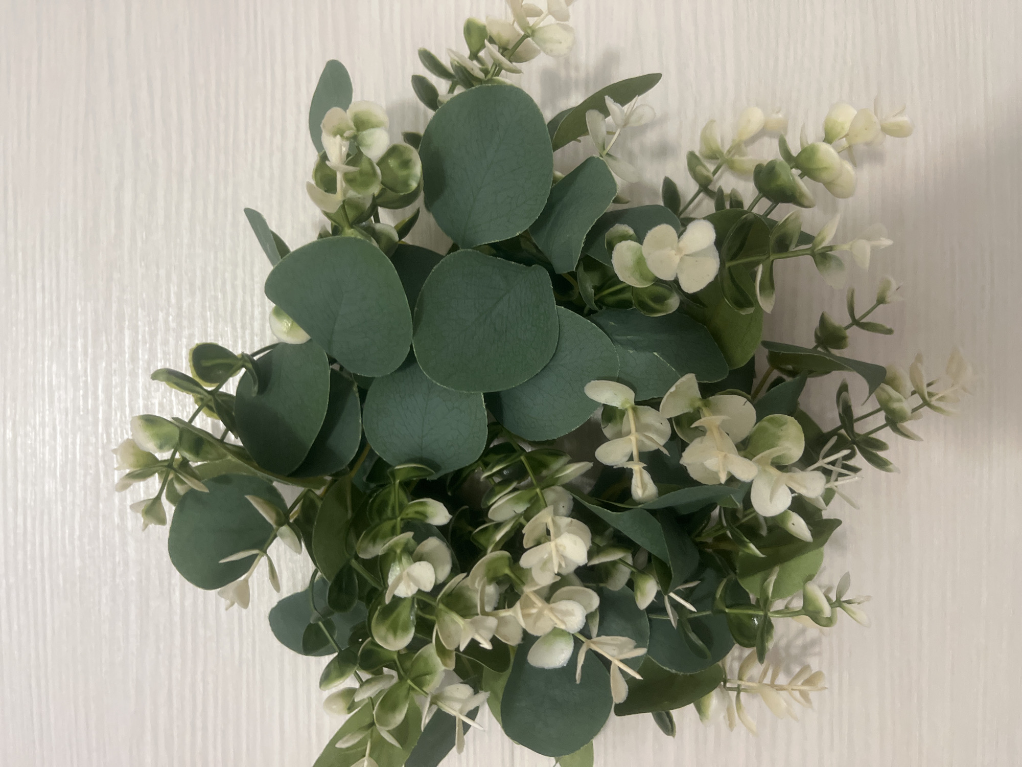 The image shows a close-up, overhead view of a floral arrangement of artificial eucalyptus branches with small white flowers. The arrangement is placed on a white, textured surface that appears to be a wooden table. There are no people or any identifiable landmarks that would indicate a location in Voronezh, Russia, or any other city. The lighting suggests an indoor setting.