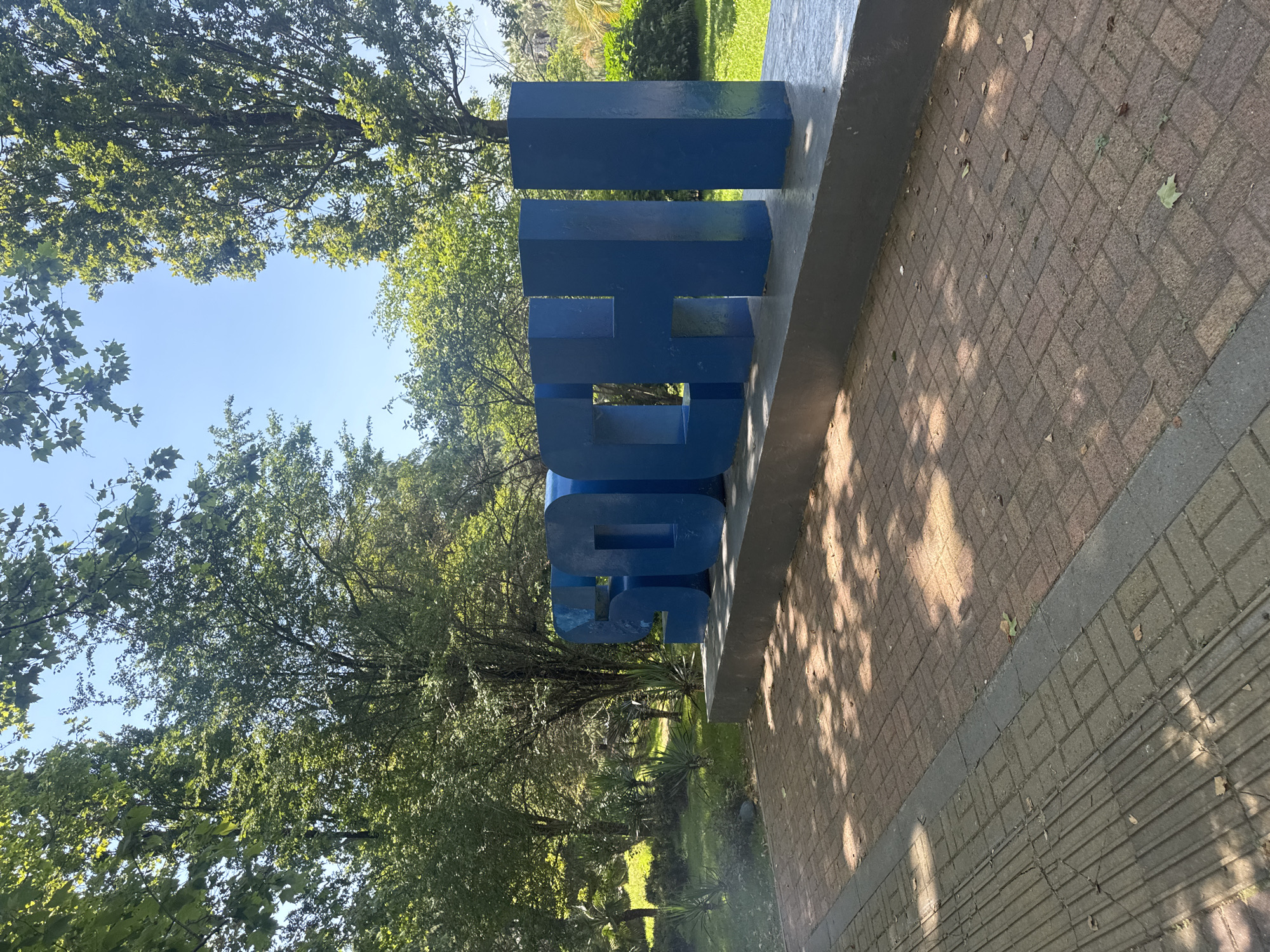 The image shows a large, blue, three-dimensional sculpture that spells out "THOS." The sculpture is positioned outdoors, with a clear blue sky visible through the branches of lush green trees. The ground in front of the sculpture is paved with reddish-brown bricks, and dappled sunlight creates shadows from the trees onto the pavement. To the right of the sculpture, the pavement transitions to a darker grey material with a textured surface. The overall scene suggests a pleasant day, likely in the late morning or early afternoon due to the bright sunlight and clear sky. The setting appears to be a park or a landscaped area, possibly on a university campus or a public space, given the monumental lettering. The sculpture is the primary object of focus, standing out against the natural backdrop of trees and sky. No people are visible in the frame. The time of day is indicated by the strong shadows and bright, even lighting. The location context of Otradnoe, Russia, does not provide further visual cues within the image itself.