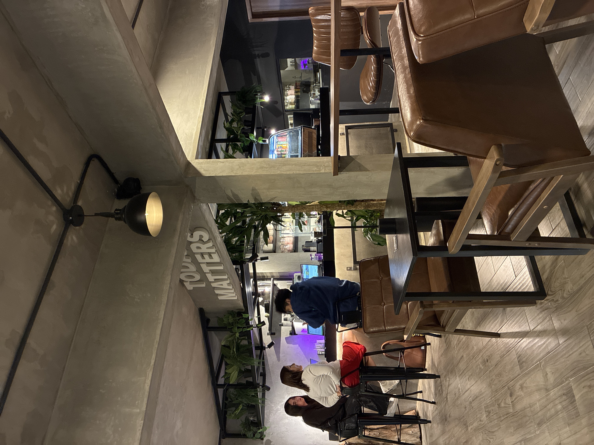 The image depicts an indoor cafe scene, likely in San Andrés Cholula, Mexico, as suggested by the prompt. The café features an industrial-chic design with exposed concrete walls and beams, highlighted by a black conduit holding a spotlight. A vertical sign reading "TODAY MATTERS" is mounted on a concrete pillar.
The foreground is dominated by brown leather chairs and tables, set against a light wood-patterned floor. In the midground, there are three people seated at a table. A person in a blue shirt is sitting at the table and seems to be focused on something on the table. Further away from them, two people are seated, with one wearing a white shirt and red pants. The third person is wearing a black jacket and sitting in the background.
The cafe design includes abundant greenery, with plants arranged along the walls and integrated into the architectural structure. A refrigerated display case containing beverages is visible in the background. The image has a muted color palette, emphasizing neutral tones of gray, brown, and green.