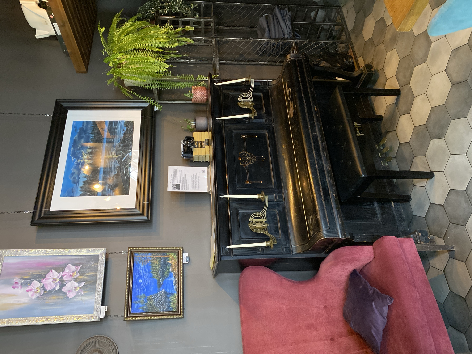 The image shows an interior scene featuring a vintage black upright piano with a bench. The piano has ornate detailing and two candlestick holders with white candles. On top of the piano, there are a few books, a vintage camera, and a small potted plant. To the left of the piano is a large, plush red armchair with a purple throw pillow. Above the piano, two framed paintings are hung on the wall, one of orchids and another of a moonlit landscape. A large framed print of a scenic natural landscape is also visible above the piano. To the right of the piano, there is a metal shelving unit with some folded clothes and a large fern plant. The floor is tiled with a hexagonal pattern of grey and white tiles. The overall setting suggests a cozy and artistic space, possibly a cafe or a private studio in Priozersk, Russia. No people are present in the image.