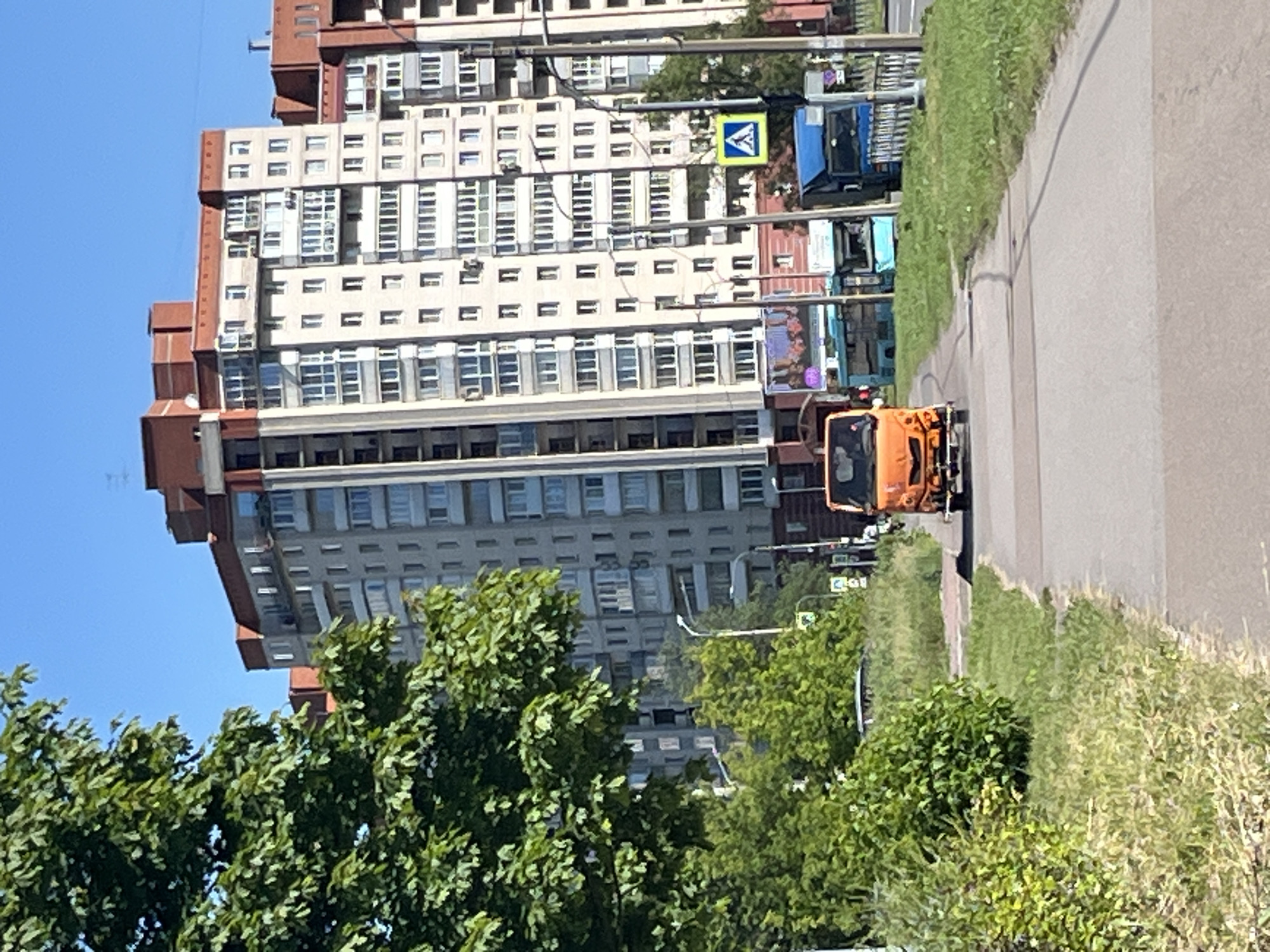 An orange utility truck is driving on a road in Saint Petersburg, Russia. The truck is in the foreground, with a sidewalk and green vegetation on either side of the road. In the background, there are tall apartment buildings. The sky is clear and blue.
