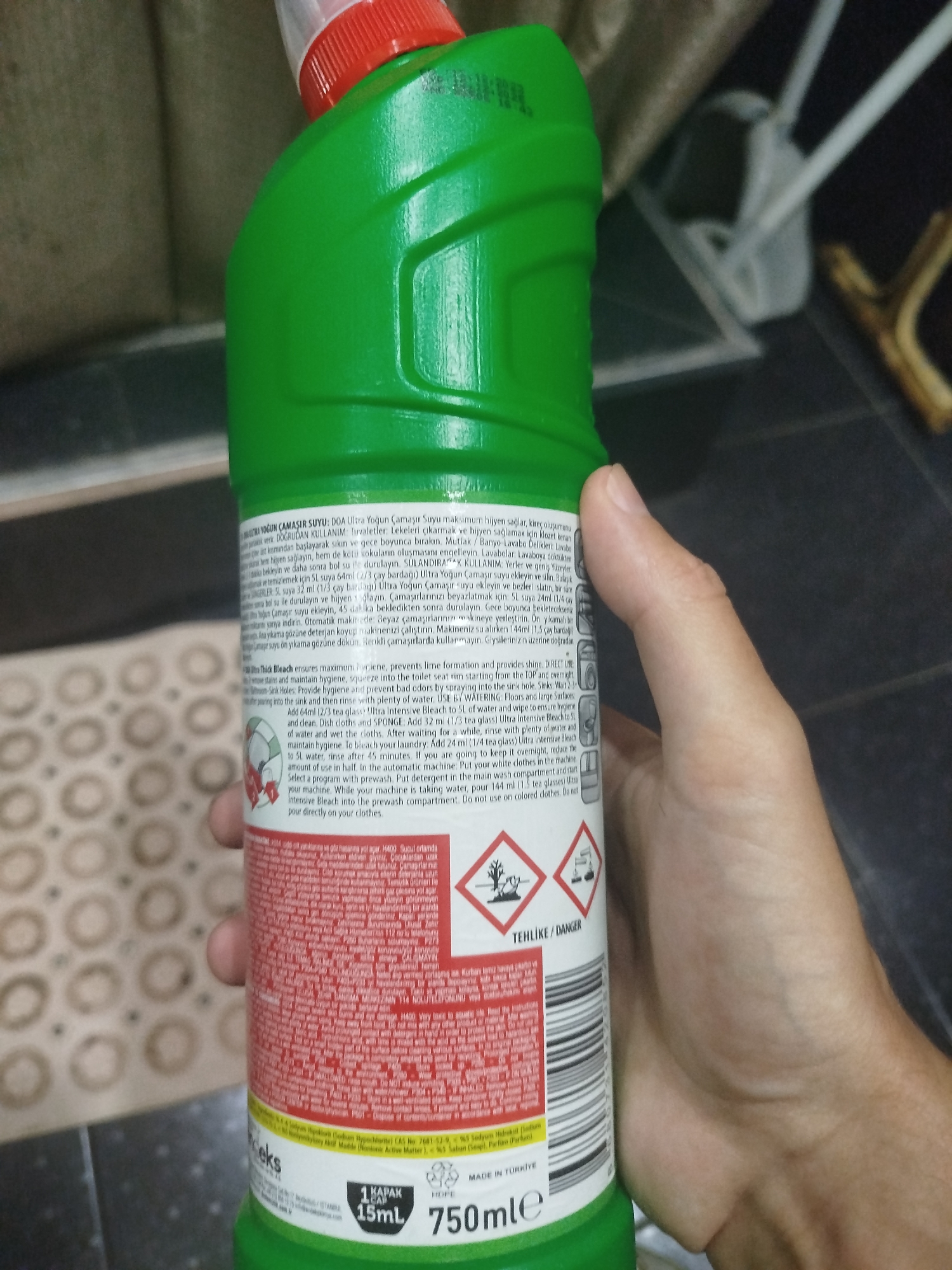 A hand holds a green plastic bottle of a cleaning product with a red cap. The bottle has a white label with text in multiple languages, including Turkish and English. The English text describes the product's uses and warnings, such as "ULTRA THICK BLEACH ensures maximum hygiene," "protect the skin," and "Do not use on colored clothes." Two hazard symbols are visible on the label: one depicting a dead tree and fish, indicating environmental danger, and another showing a hand being corroded by liquid, signifying corrosive properties. The background is blurred but appears to be indoors, possibly a domestic setting, with hints of furniture and flooring. The image is taken from a slightly low angle, with the bottle positioned vertically in the foreground. There is no indication of the city of Batumi, Georgia, or any specific urban environment.