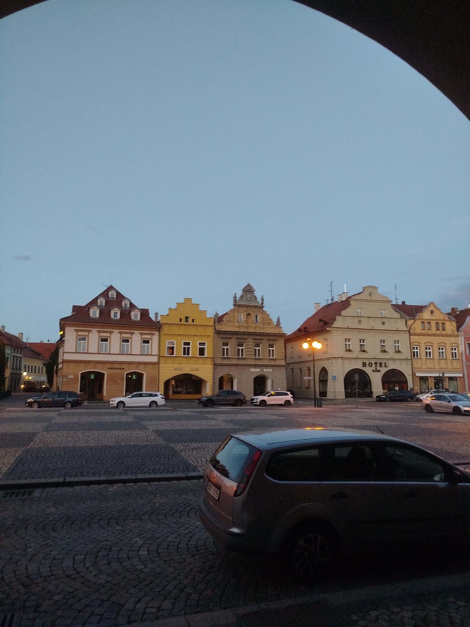 An eye-level, vertical shot captures a street scene at dusk in a European city, likely the Czech Republic given the architectural style. The sky is a muted blue, transitioning to darker tones near the top, with a dark archway framing the upper portion of the image.
The focal point is a row of traditional, multi-story buildings lining the street. From left to right, the first building is a light brown and white structure with a red tiled roof and multiple dormer windows. It features arched entrances on the ground floor, one with a sign that appears to read "LÉKÁRNA" (pharmacy). Next to it is a bright yellow building with a stepped gable roof and arched ground floor openings, housing shops. Further right, a pale yellow building with a red tiled roof is prominent, featuring a large arched entrance with the word "HOTEL" clearly visible in dark lettering above it. Adjacent to the hotel, more buildings in shades of yellow and pink complete the row, showing similar architectural elements with pitched roofs and dormer windows.
The street itself is paved with cobblestones arranged in a radial pattern. Several cars are parked or driving on the street. A dark gray sedan is parked on the left. A white SUV is driving in the middle of the street. Another dark gray sedan is parked further to the right, and a silver sedan is seen in the distance. In the foreground, the rear of a silver hatchback car is visible, taking up the lower right quadrant of the image. A lamppost with a warm orange glow stands near the hotel, providing illumination. The overall impression is a quiet, historic urban setting at the end of the day.