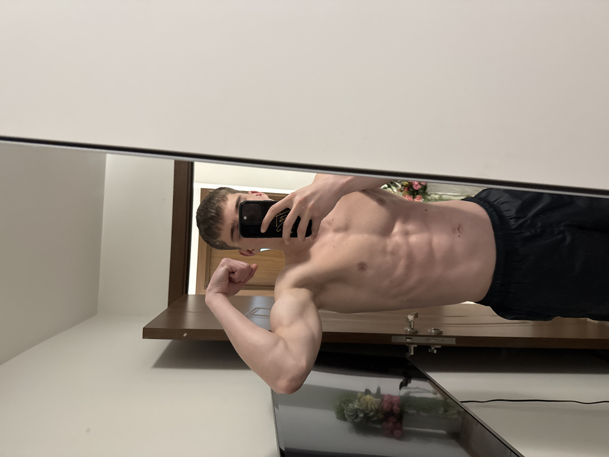 A young man takes a mirror selfie in an indoor setting in Varna, Bulgaria. He is shirtless, revealing a muscular physique with prominent abdominal definition. He wears dark shorts and has short, dark hair. With his right hand, he holds a dark-cased smartphone to capture his reflection, while his left arm is flexed, showcasing his bicep.
The background reflected in the mirror features a light-colored wall, a wooden door, and a brown shelf or piece of furniture. A subtle reflection of what appears to be a colorful floral or plant arrangement can be seen on a surface below the mirror's frame. The lighting suggests an indoor environment, possibly with artificial light. The smartphone case has a golden design or text, which is not clearly legible. The overall scene depicts a posed, confident stance.
