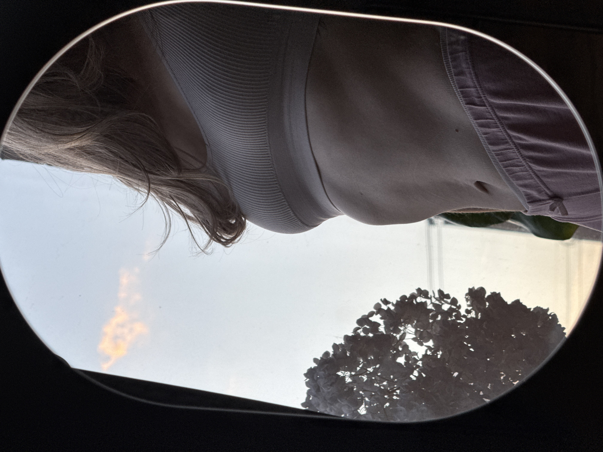 A person wearing a ribbed tank top and drawstring pants is looking down through an open sunroof, likely of a car, showing the sky and silhouetted tree branches outside. The location is not discernible from the image.